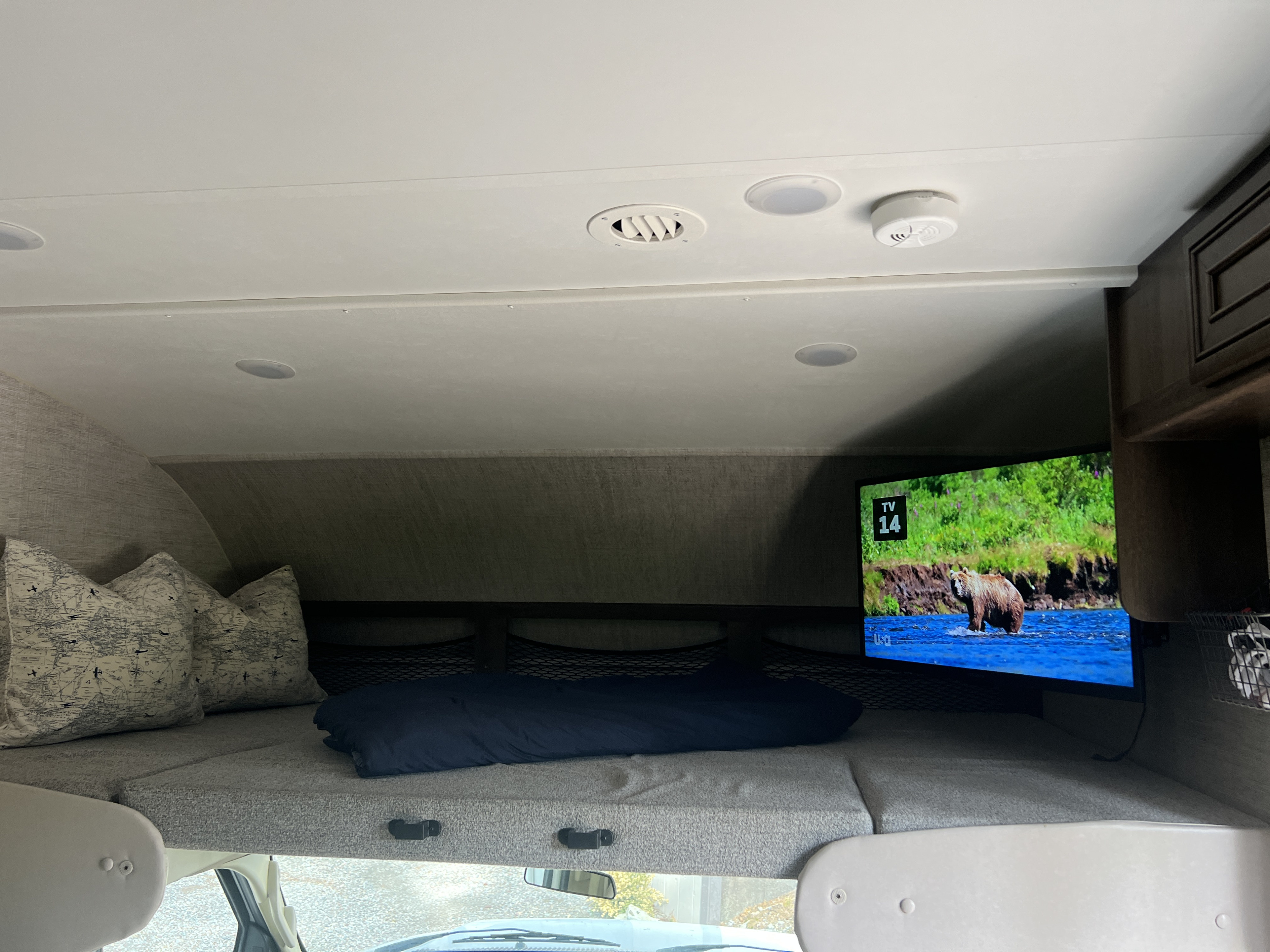 Cozy RV rental cab-over bunk features a TV showing a bear, overhead lights, and a smoke detector, providing a comfortable sleeping area.. Jayco Redhawk 2021