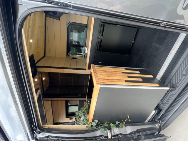 Modern RV rental interior, viewed from the rear. Features wood paneling, compact kitchen area with sink, and mounted TV.. Ford Transit Custom 2023