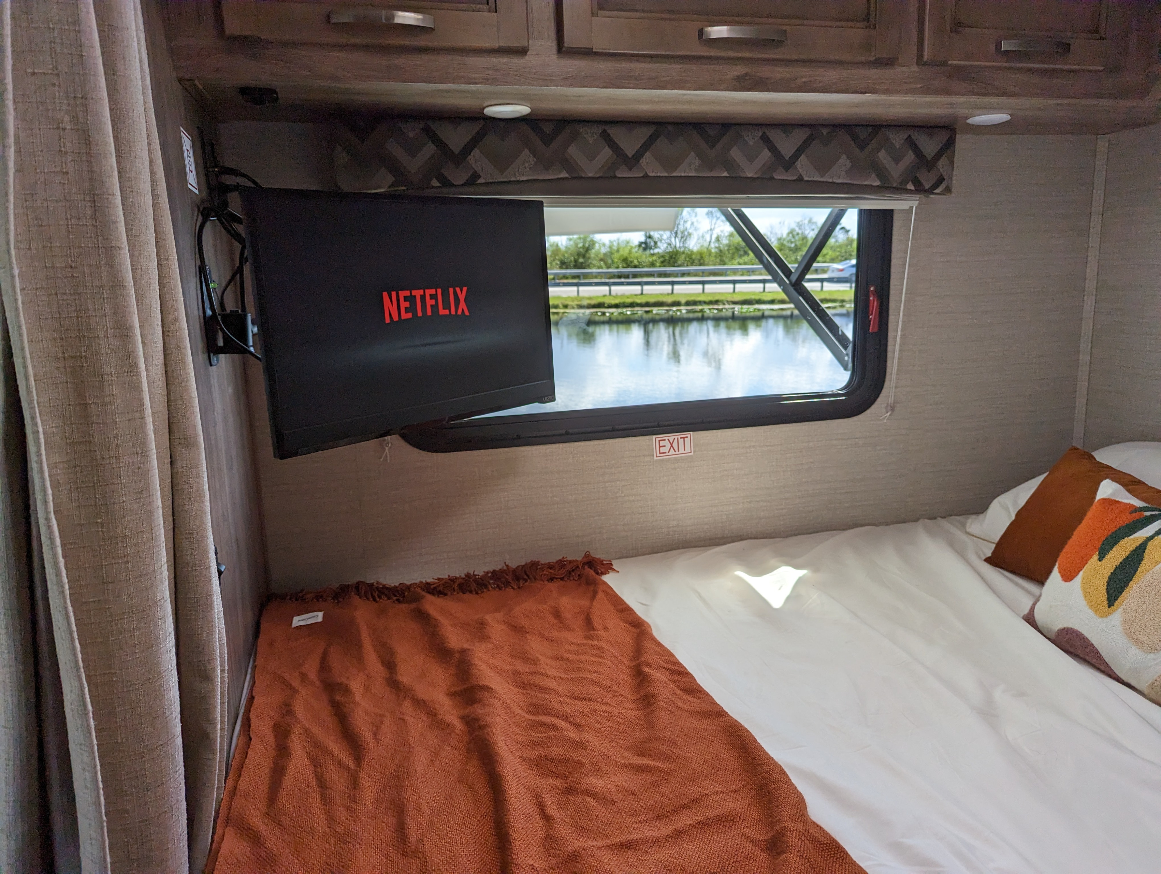 Cozy rv rental bedroom: A mounted TV with Netflix, a bed with an orange throw, and a window view of water & road.. Jayco Redhawk SE 2020