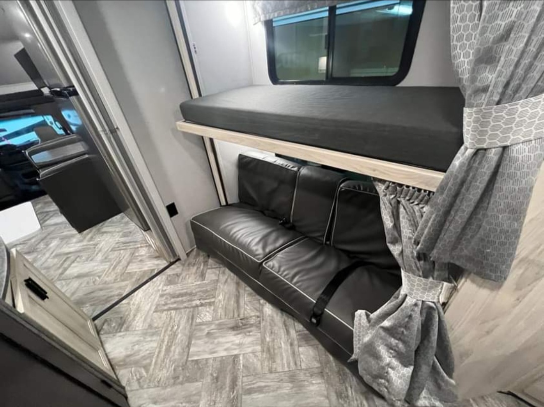 Cozy RV rental interior with a bunk bed over a black sofa, complete with seatbelts, patterned curtains, and wood-look floor.. Forester LE 2022