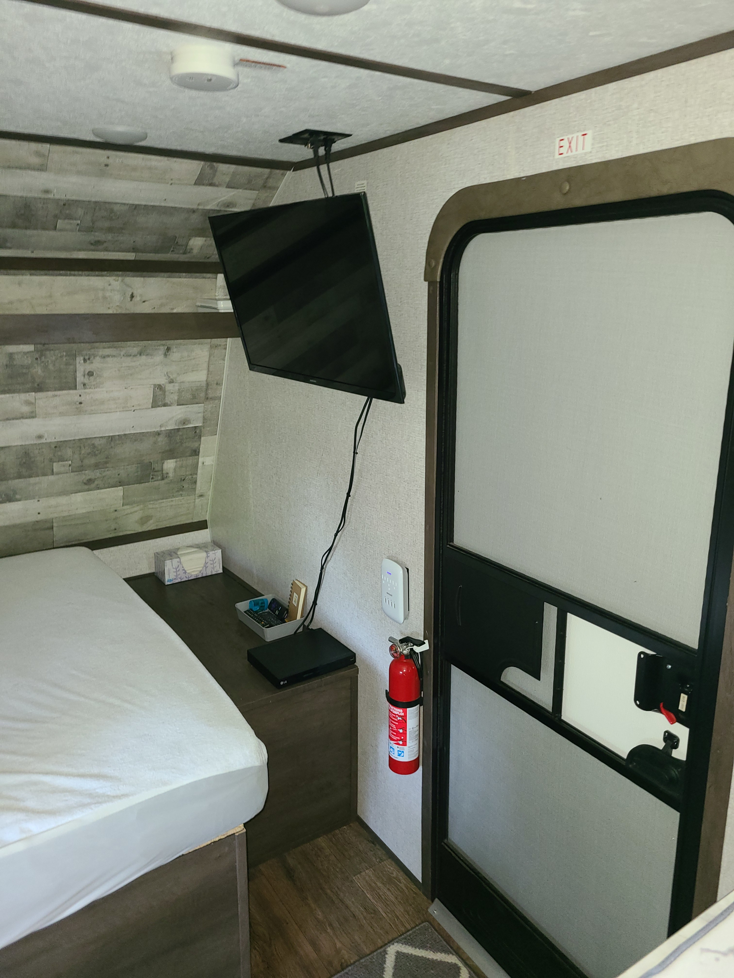 Interior of an rv rental with a bed, wall-mounted TV, and a fire extinguisher near the main door. An 'EXIT' sign is visible.. Dutchmen Coleman Lantern LT 2021