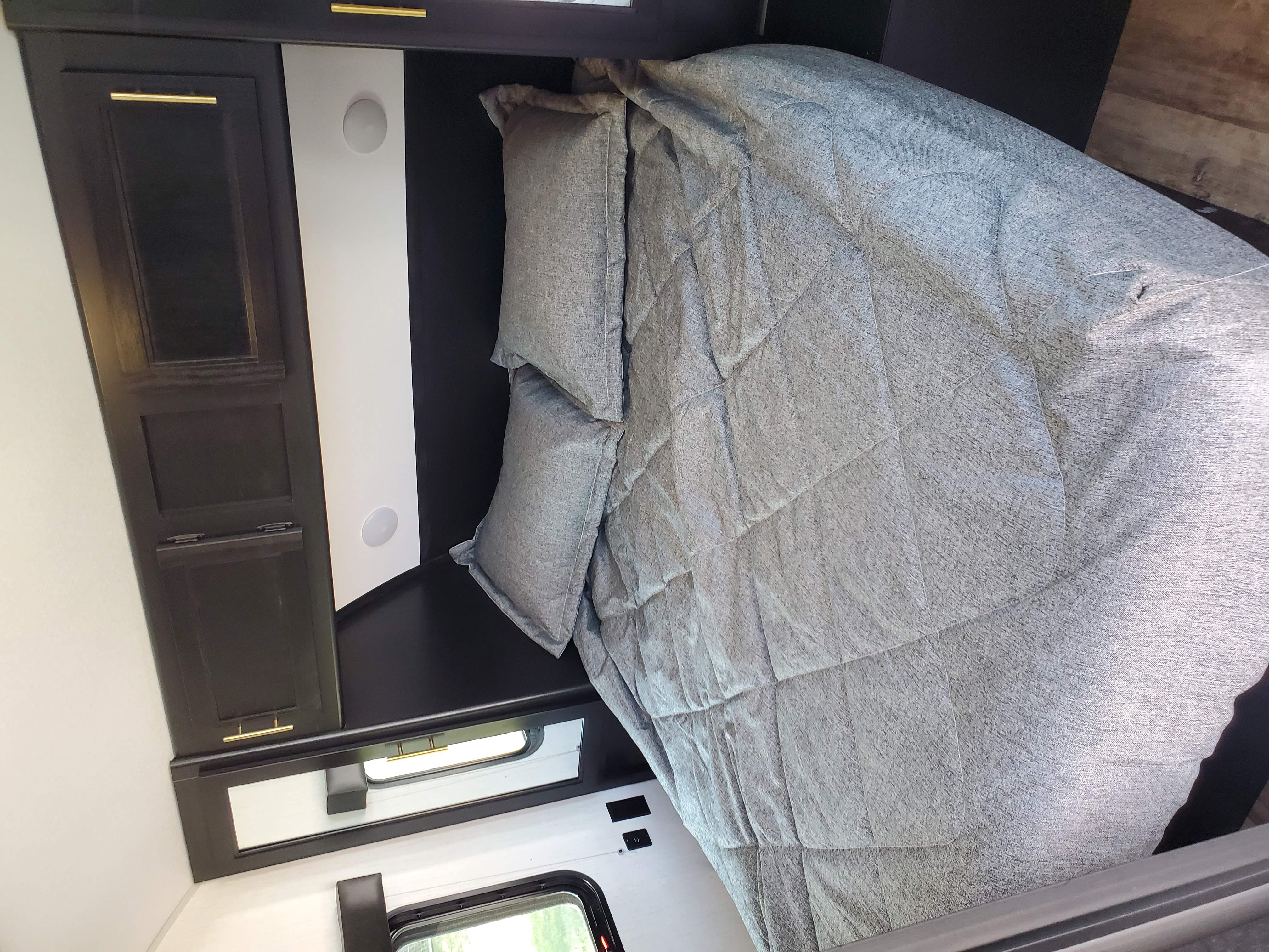 Cozy RV rental bedroom with grey comforter, pillows, dark wood cabinets, and a window. A white circular fixture is also visible.. Forest River Cherokee 2022