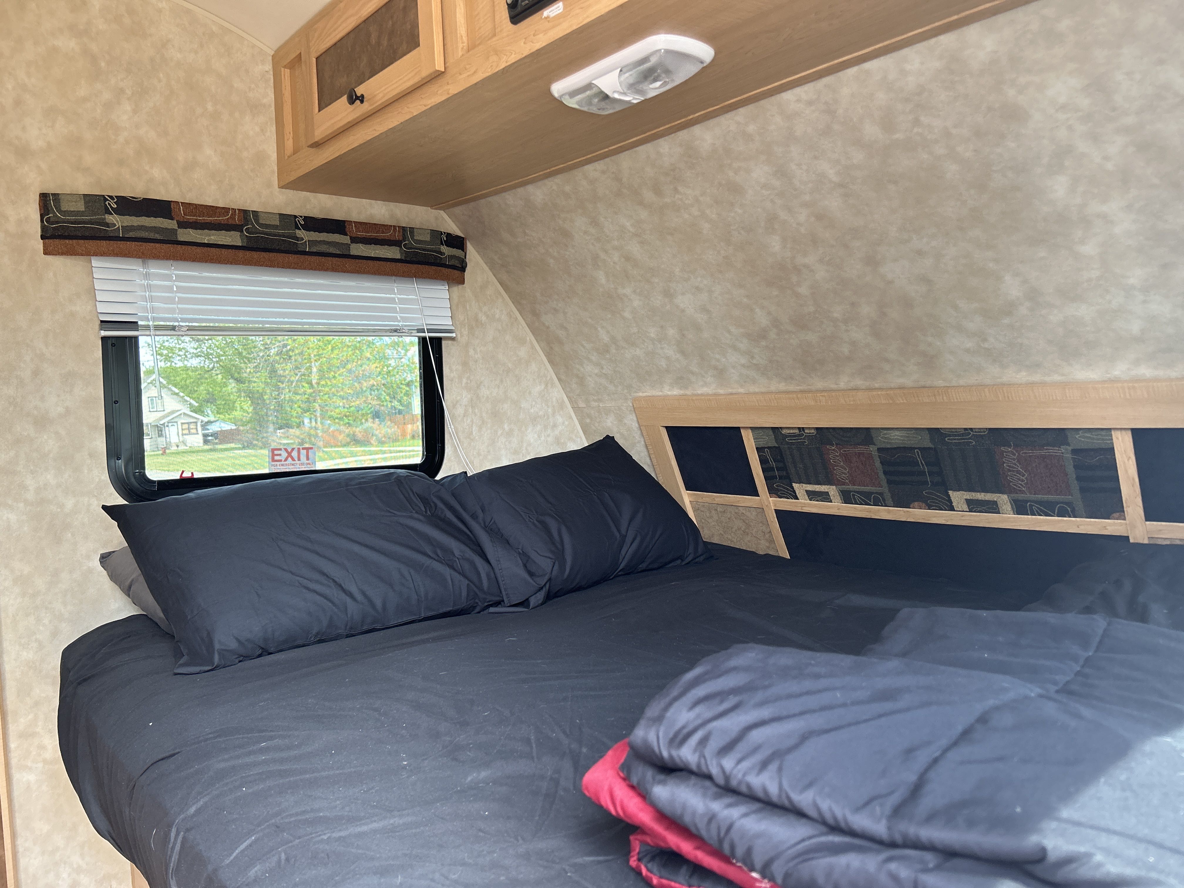 Relaxing RV rental bedroom setup with black bedding, window showing a house, overhead cabinets, and an "EXIT" sign.. Ez Coyote lite 2010