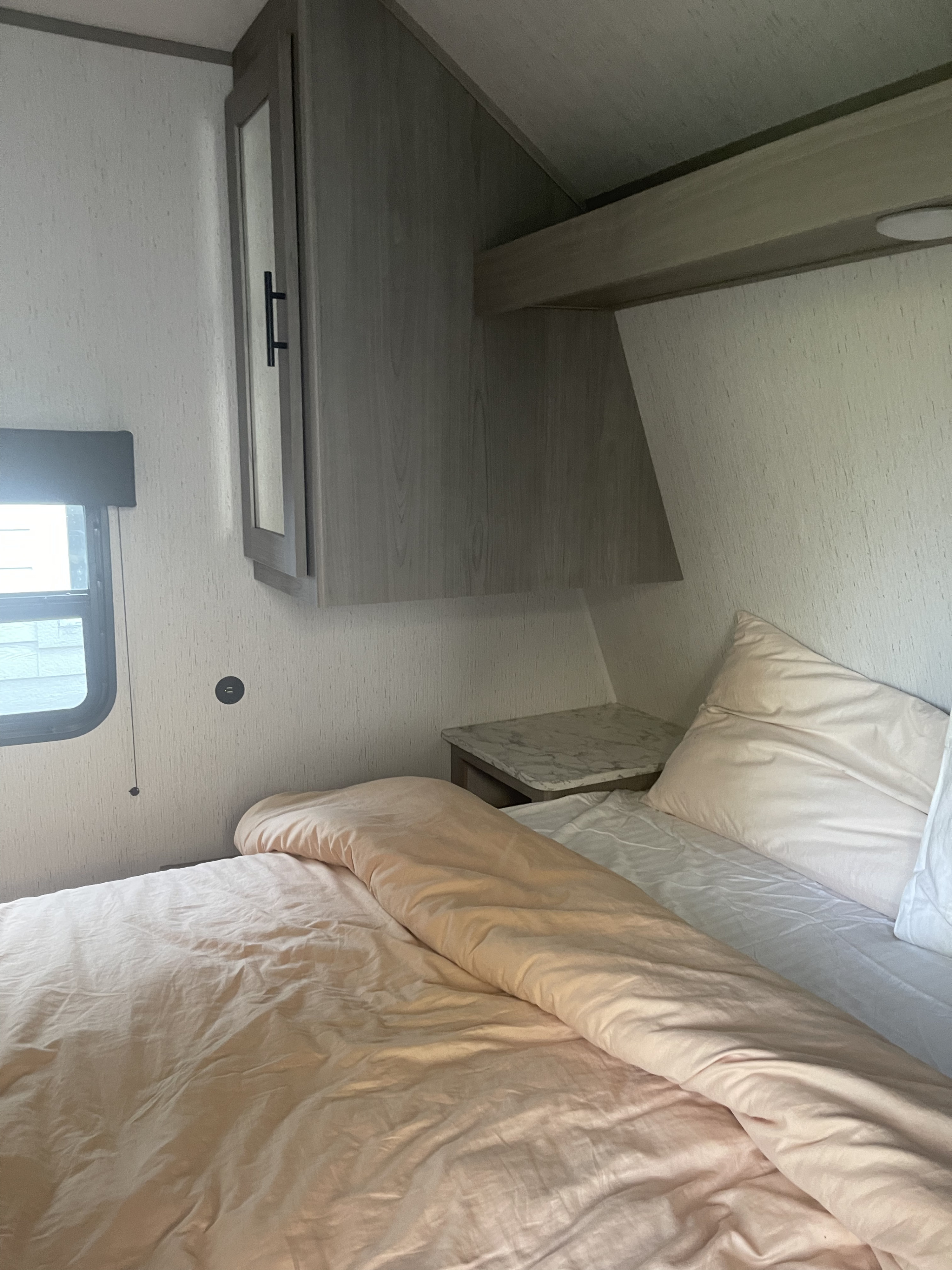 Cozy RV rental bedroom featuring a bed with peach bedding, overhead cabinet, window, and a convenient USB outlet.. Coleman Dutchman Lantern 2022