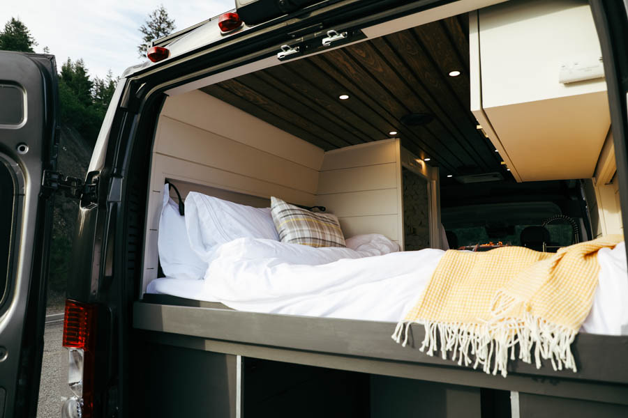 Cozy, white-sheeted bed with a yellow throw inside an RV rental camper van, seen from the open rear doors, hinting at comfortable mobile living.. Other Other 2022