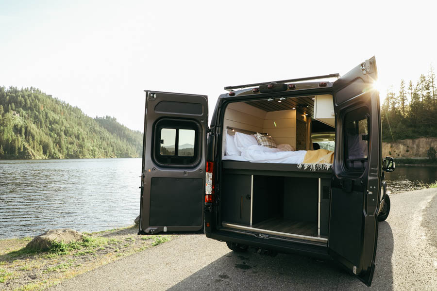 RV rental parked lakeside. Open doors reveal a cozy bed & storage inside, offering scenic views for travel.. Other Other 2022