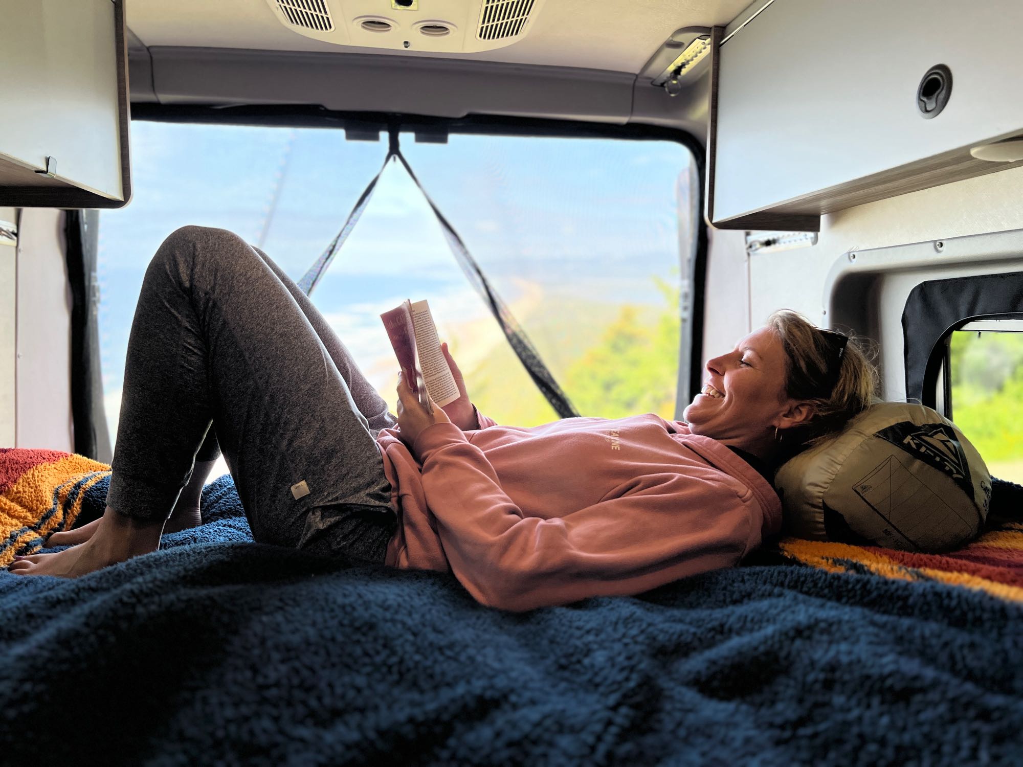 A woman smiles while relaxing and reading a book inside a cozy RV rental, with a scenic outdoor view visible through the rear window.. Winnebago Solis 2023