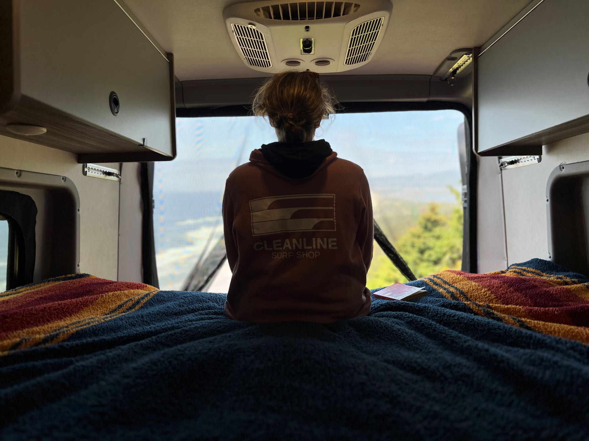 Inside a comfy RV rental, a person looks out the back window at a scenic view from their bed. Overhead cabinets are visible.. Winnebago Solis 2023