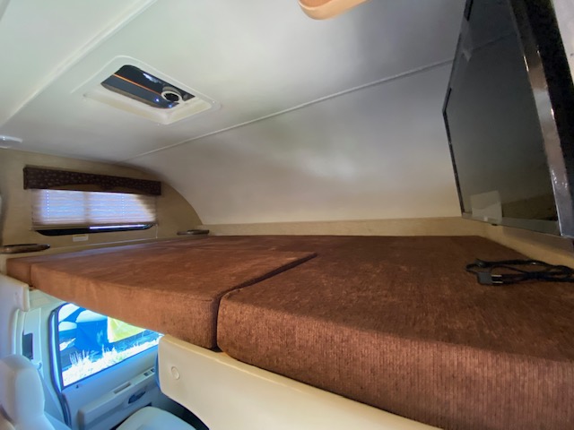 An RV rental's cab-over bunk space, featuring a brown cushioned bed, side window, ceiling vent, and mounted TV.. Thor Chateau 2015