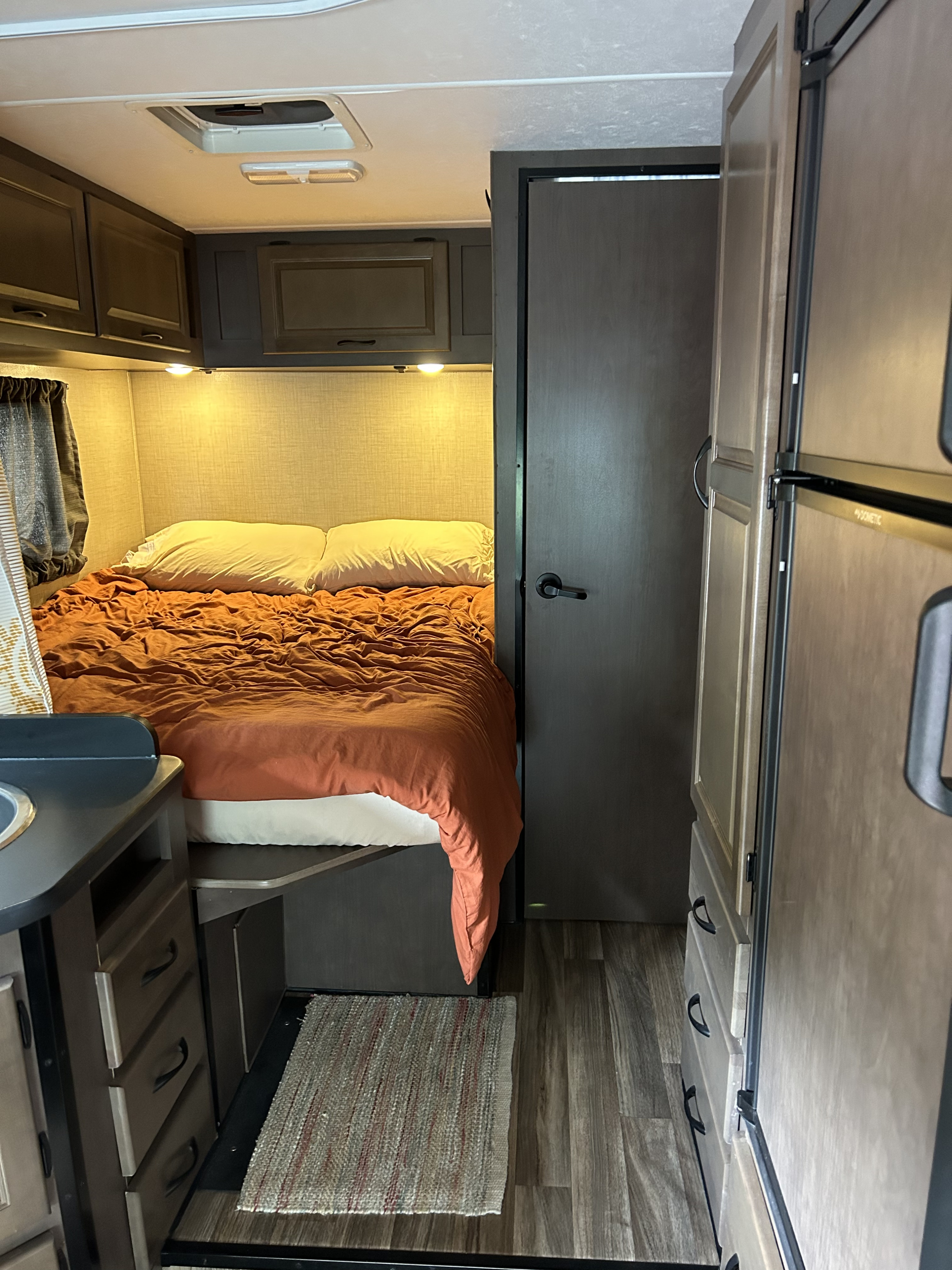 Cozy RV rental interior with compact bedroom featuring orange bedding, small kitchenette, and refrigerator.. Thor Majestic 2017