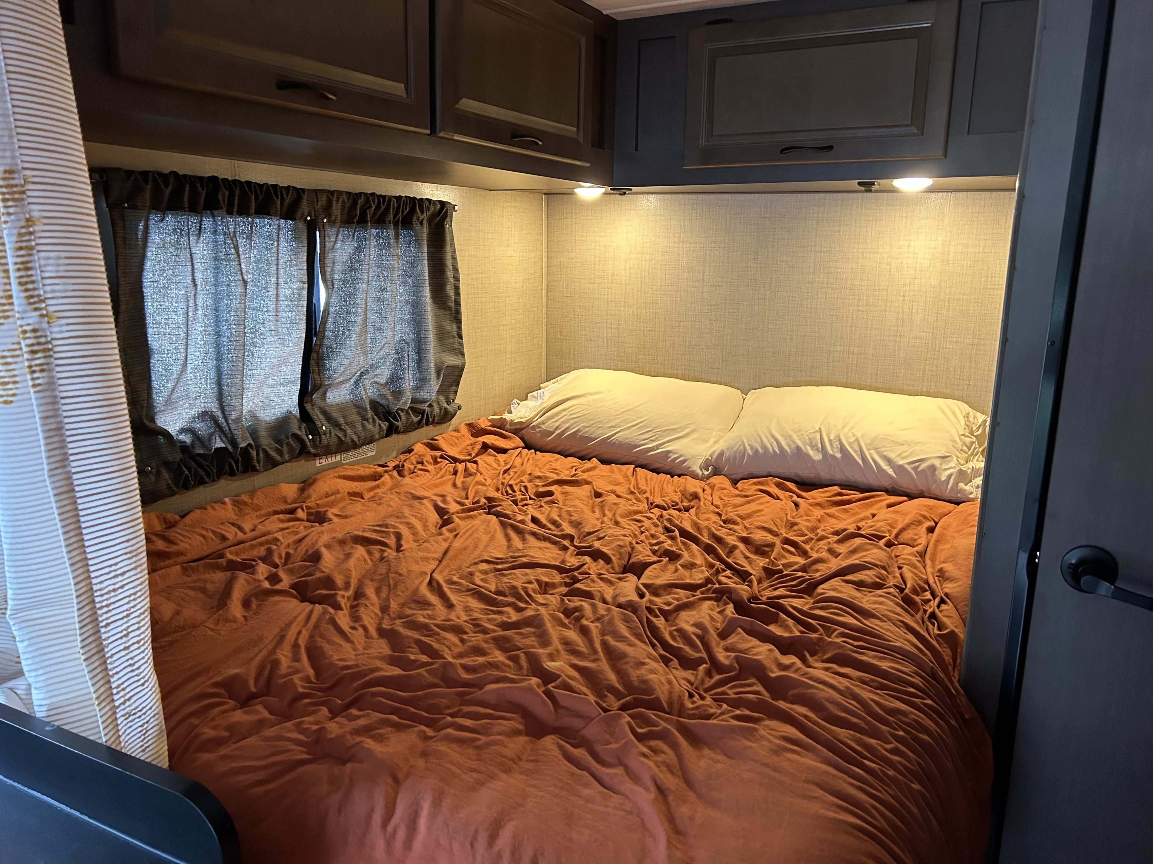 Inside a cozy RV rental bedroom: an inviting bed with rust-colored linens, white pillows, cabinets, and warm lighting.. Thor Majestic 2017