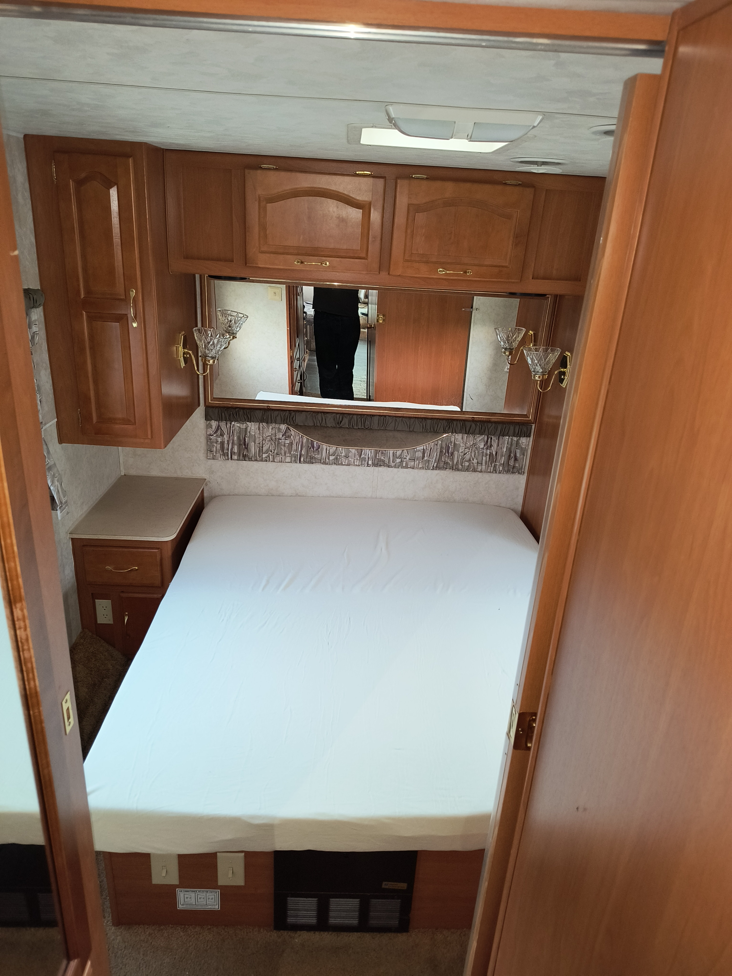 RV rental bedroom with a clean white bed, wooden overhead cabinets, a mirror, and a built-in nightstand.. Damon Corporation Daybreak 2001