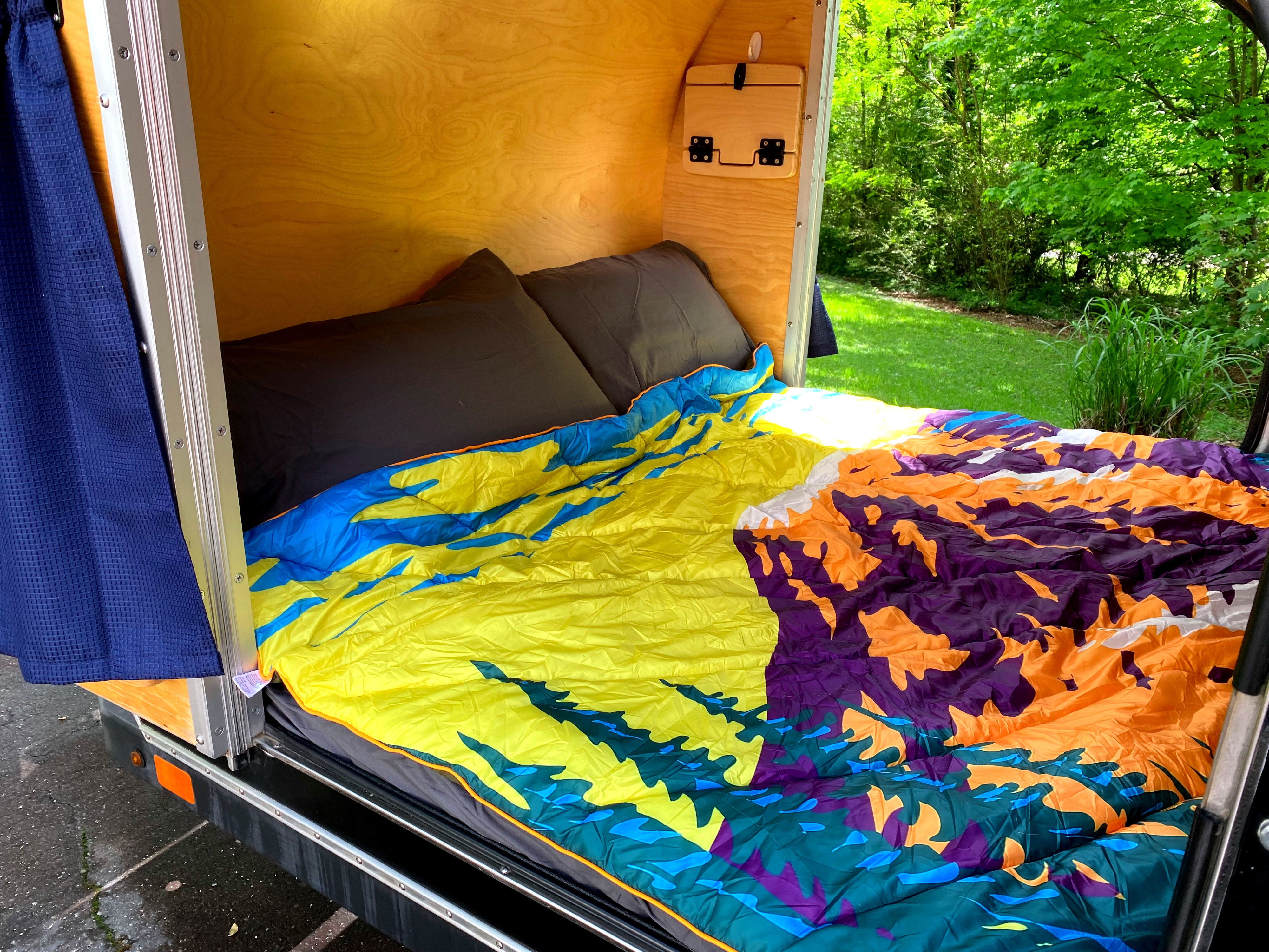 Colorful bedding and dark pillows in a cozy, wood-paneled RV rental bedroom, with a view of green trees outside.. SoCal Krawler 459 2011