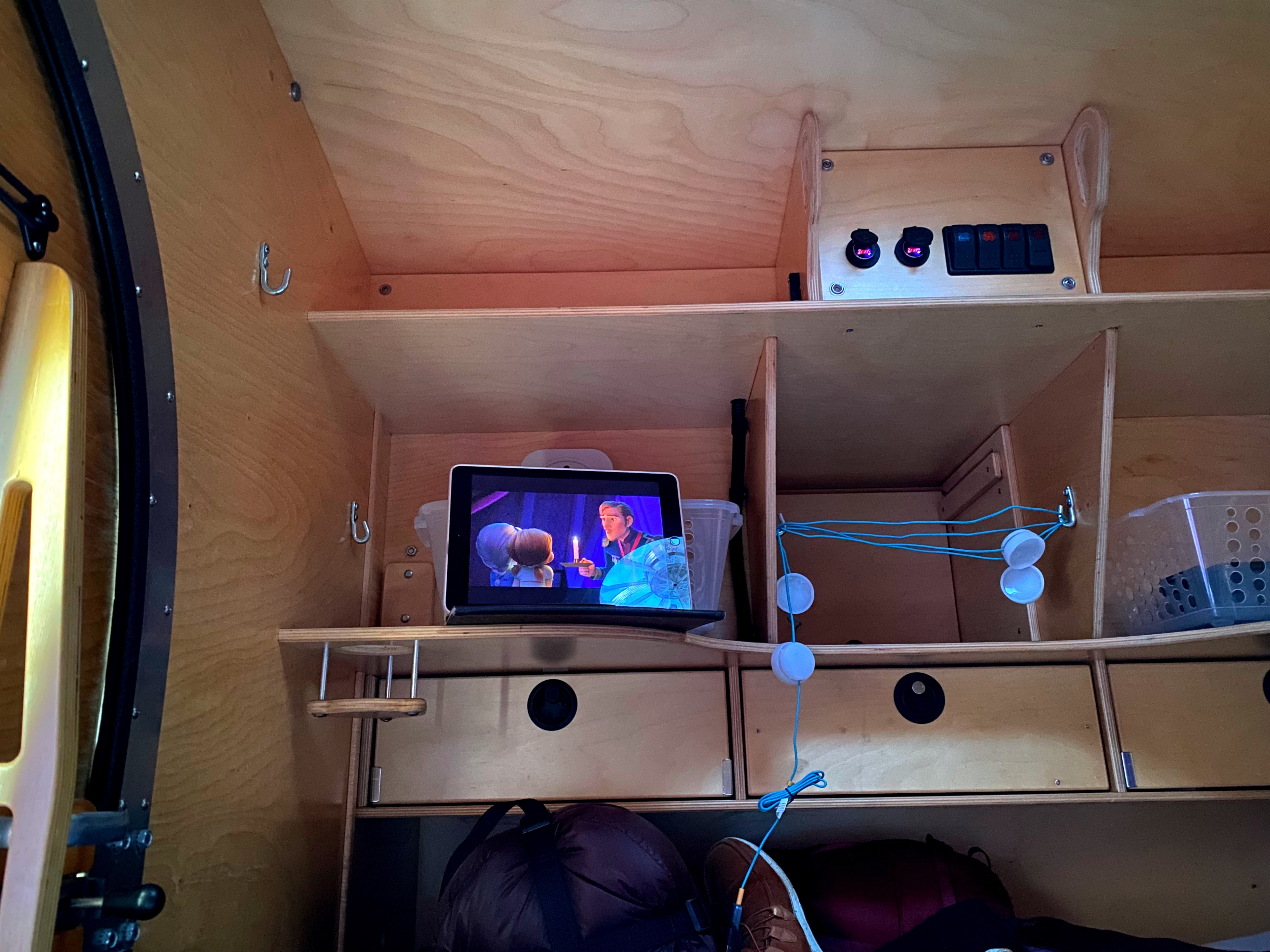 Inside an RV rental: a compact, wooden interior featuring shelves, drawers, and a tablet playing a movie. Charging ports visible.. SoCal Krawler 459 2011