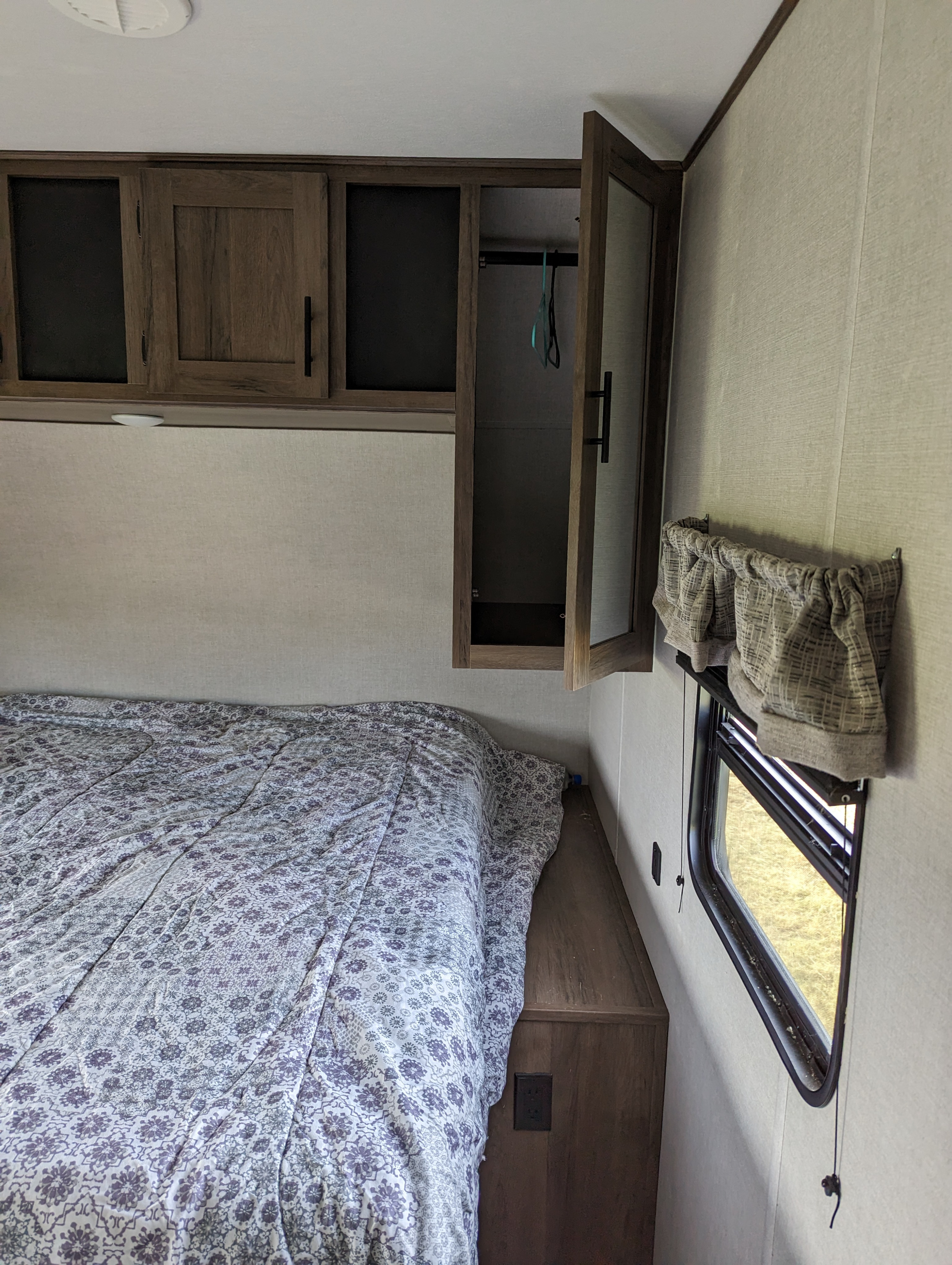 Inside an RV rental bedroom: features a patterned bed, wood overhead cabinets (one open), and a window with blinds.. Primtetime Avenger 2021