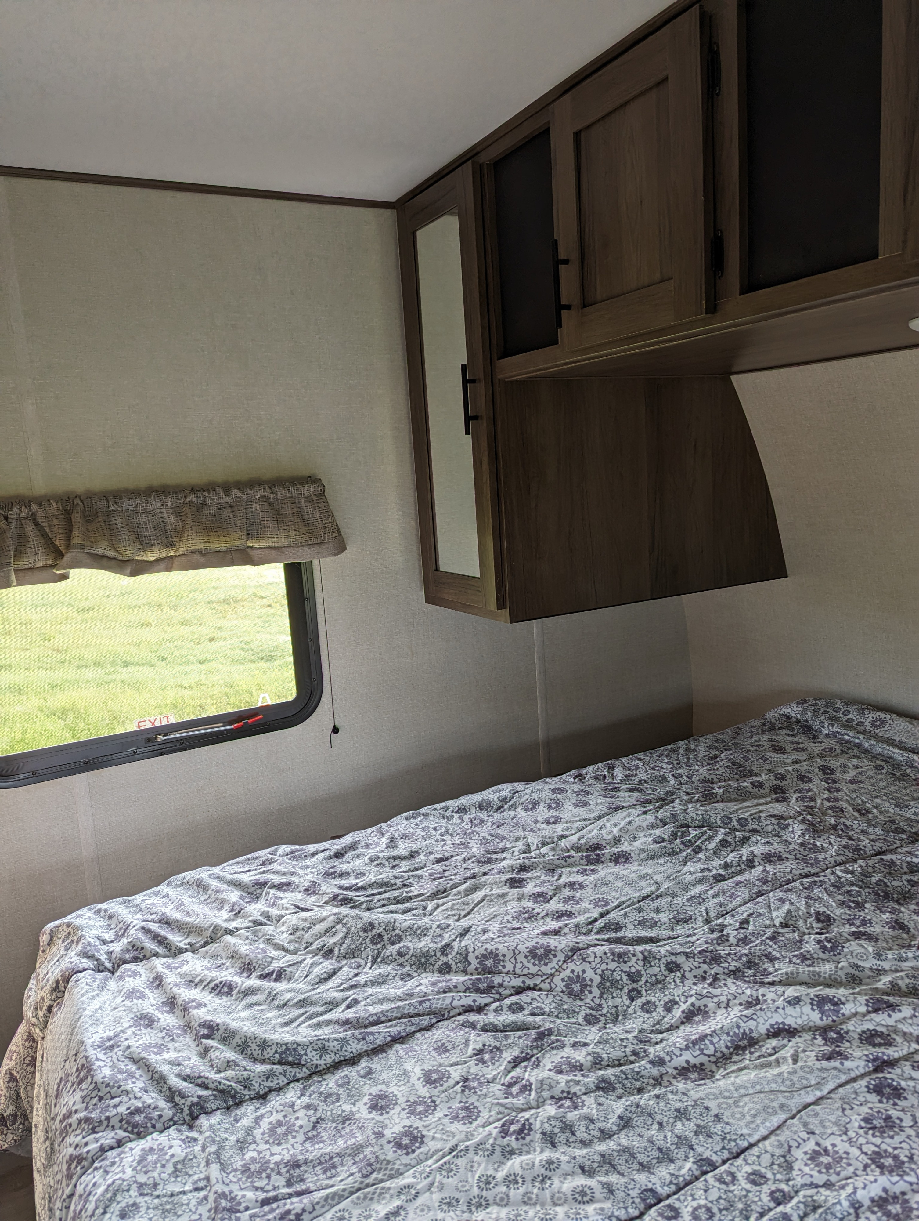 Cozy RV rental bedroom with a patterned bed, overhead cabinets, and a window looking out onto a green field.. Primtetime Avenger 2021