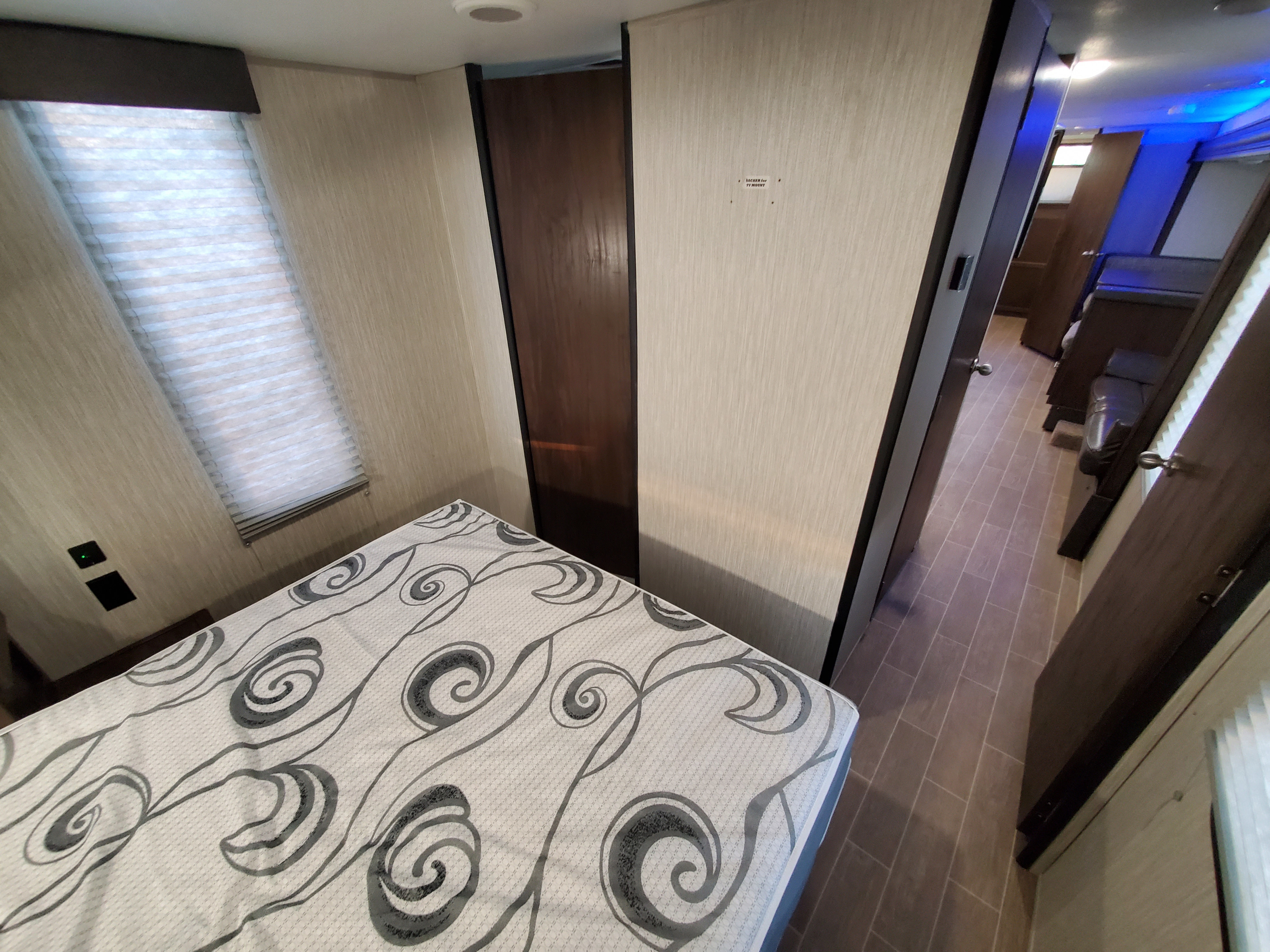 Modern RV rental bedroom with patterned mattress, pleated window, and hallway view. Wall outlets visible.. Forest River patriot 2020