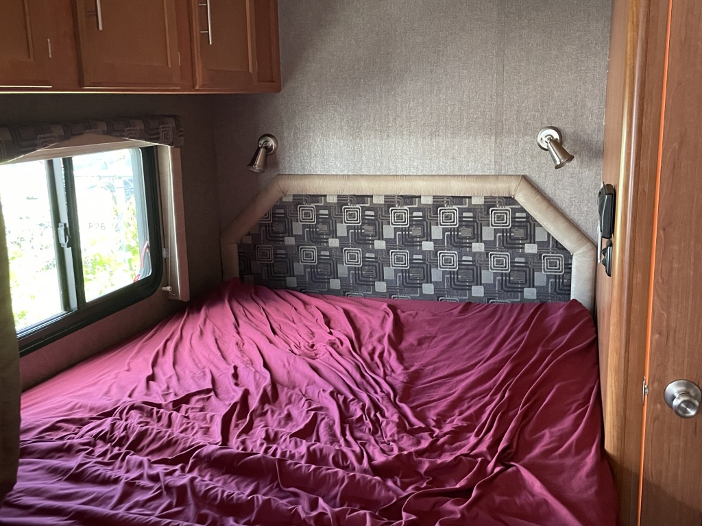 Experience an RV rental: bedroom with red-covered bed, patterned headboard, window, and overhead cabinets.. Adventurer Adventurer Motorhome 2017