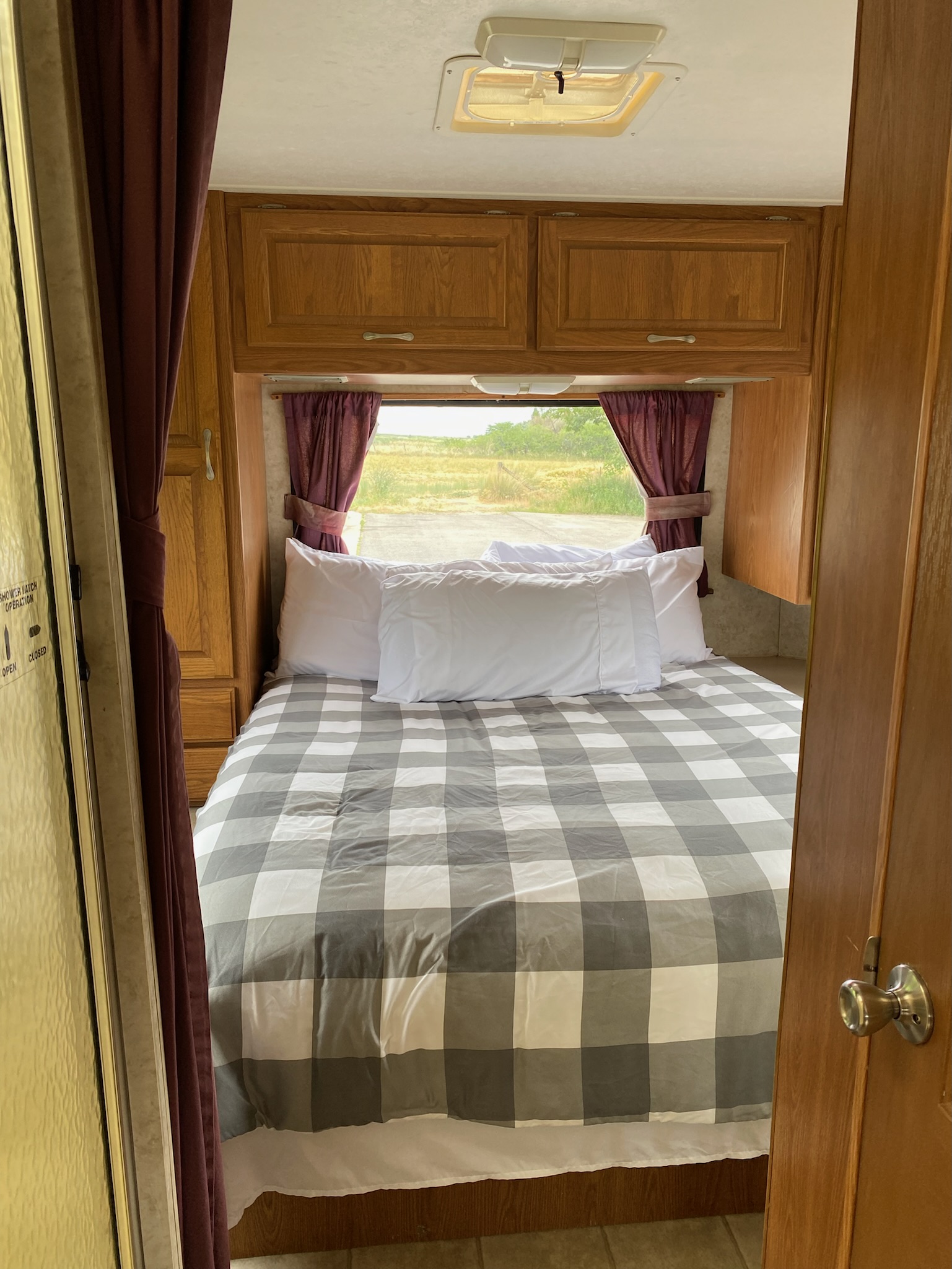 Cozy RV rental bedroom with comfortable gray plaid bedding, overhead wooden cabinets, and a bright window view.. Four Winds Majestic 2004