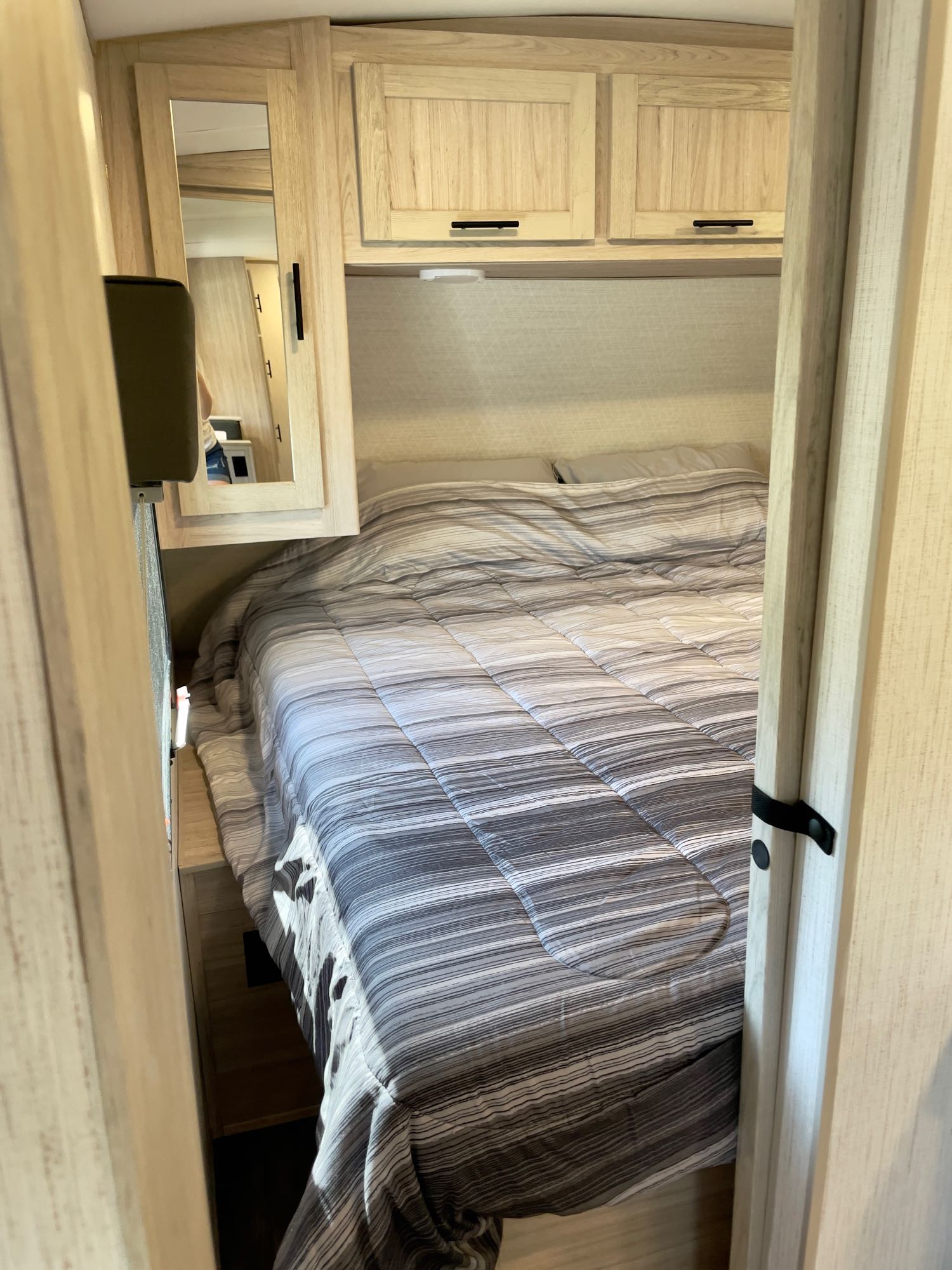 Inside an RV rental bedroom: a striped bed, light wood overhead cabinets, and a full-length mirror showing a reflection.. Cruiser Rv Corp Shadow Cruiser 2023