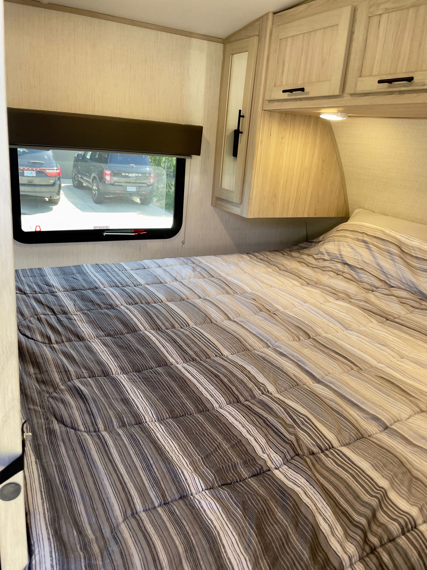 Inside an RV rental: a bedroom with a striped comforter (brown/grey/white), overhead cabinets, and window showing cars.. Cruiser Rv Corp Shadow Cruiser 2023