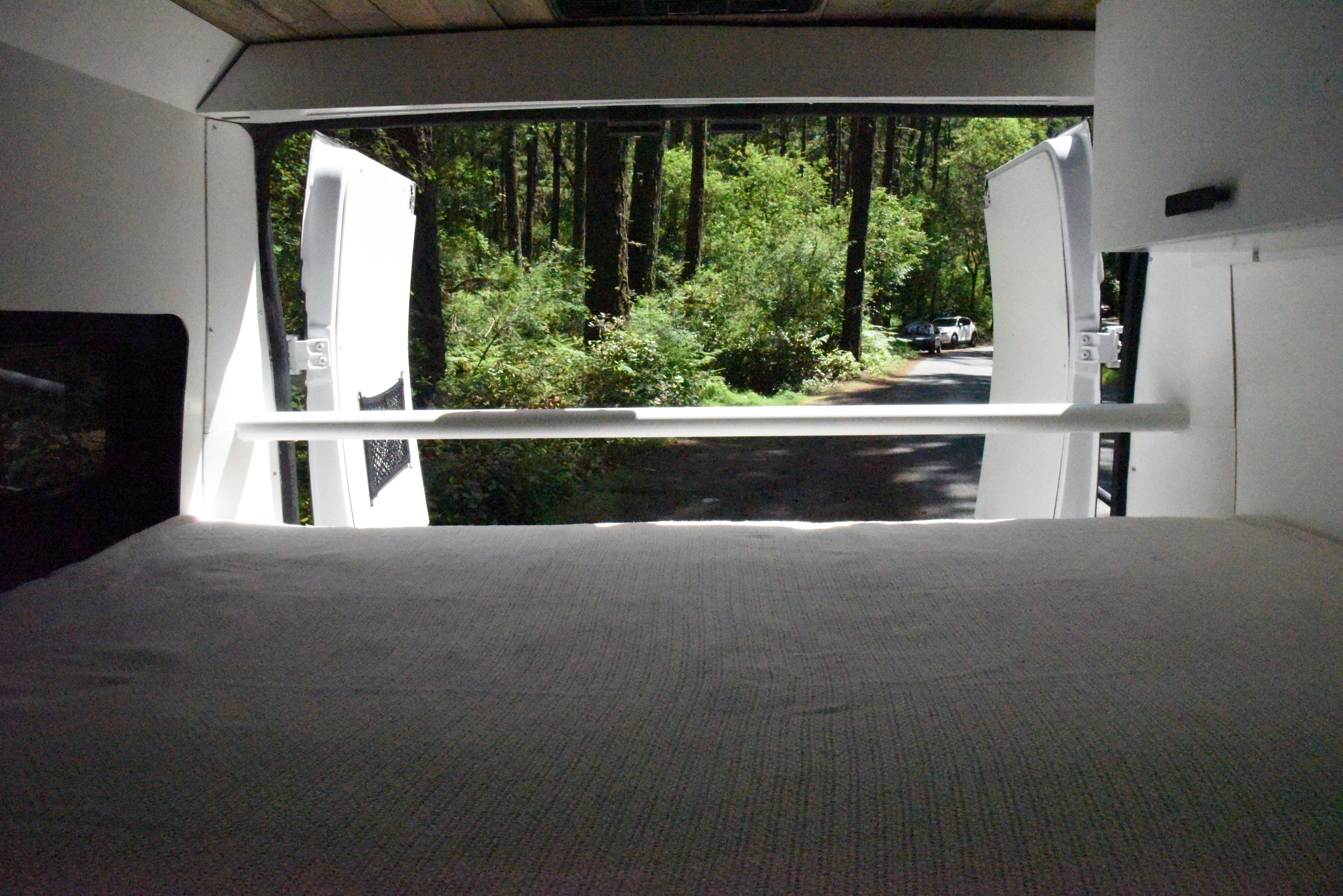 An RV rental interior, from the bed, showing open back doors leading to a vibrant forest road with parked cars. Ready for adventure!. Ram Promaster 2019