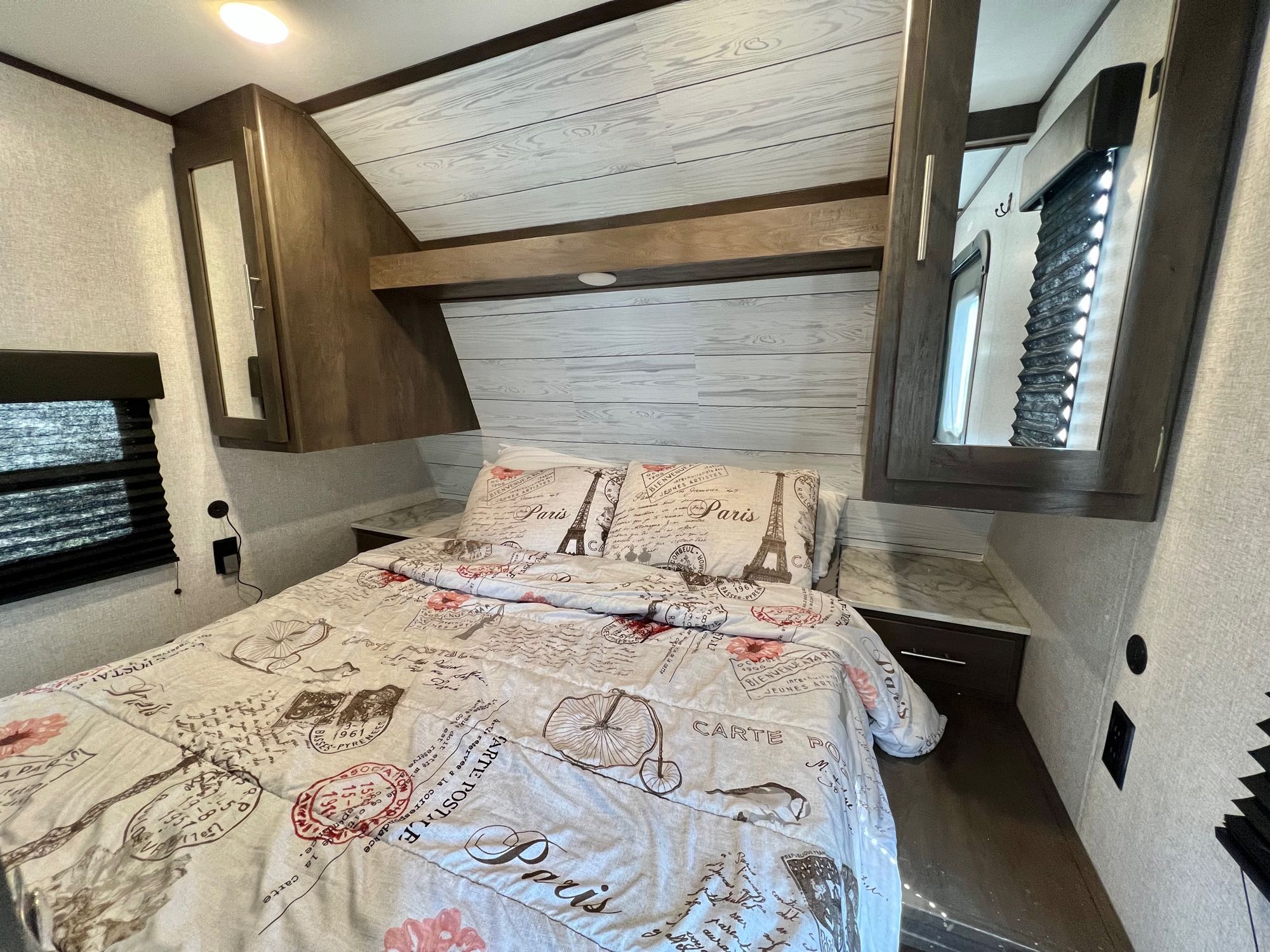 Cozy RV rental bedroom interior, featuring Paris-themed bedding, overhead cabinets, and a window with blinds.. Dutchmen Coleman 2021
