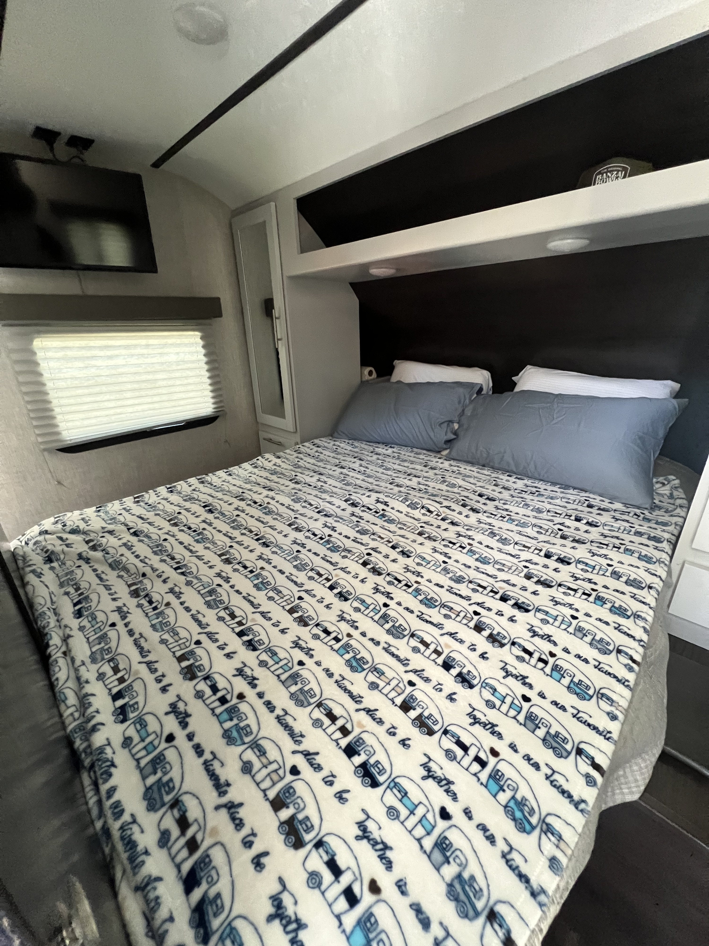 Cozy RV rental bedroom with a bed featuring a camper-patterned blanket, blue pillows, wall-mounted TV, and overhead storage.. Keystone RV Bullet Ultra Lite 2020
