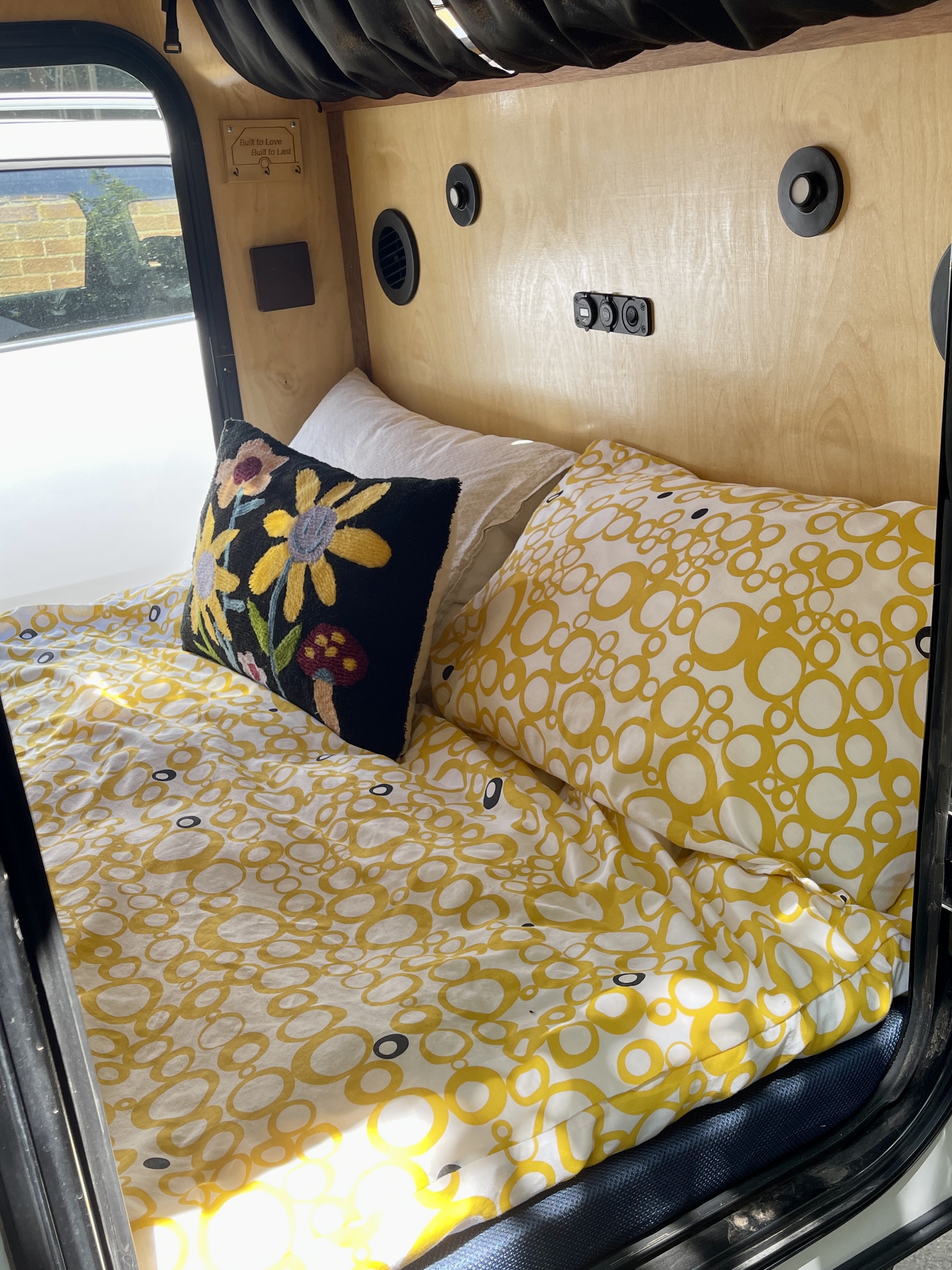 Inside this cozy rv rental, a bed with cheerful yellow and white patterned bedding, a floral pillow, and wood walls.. Vintage Trailer Works XTR Other 2023