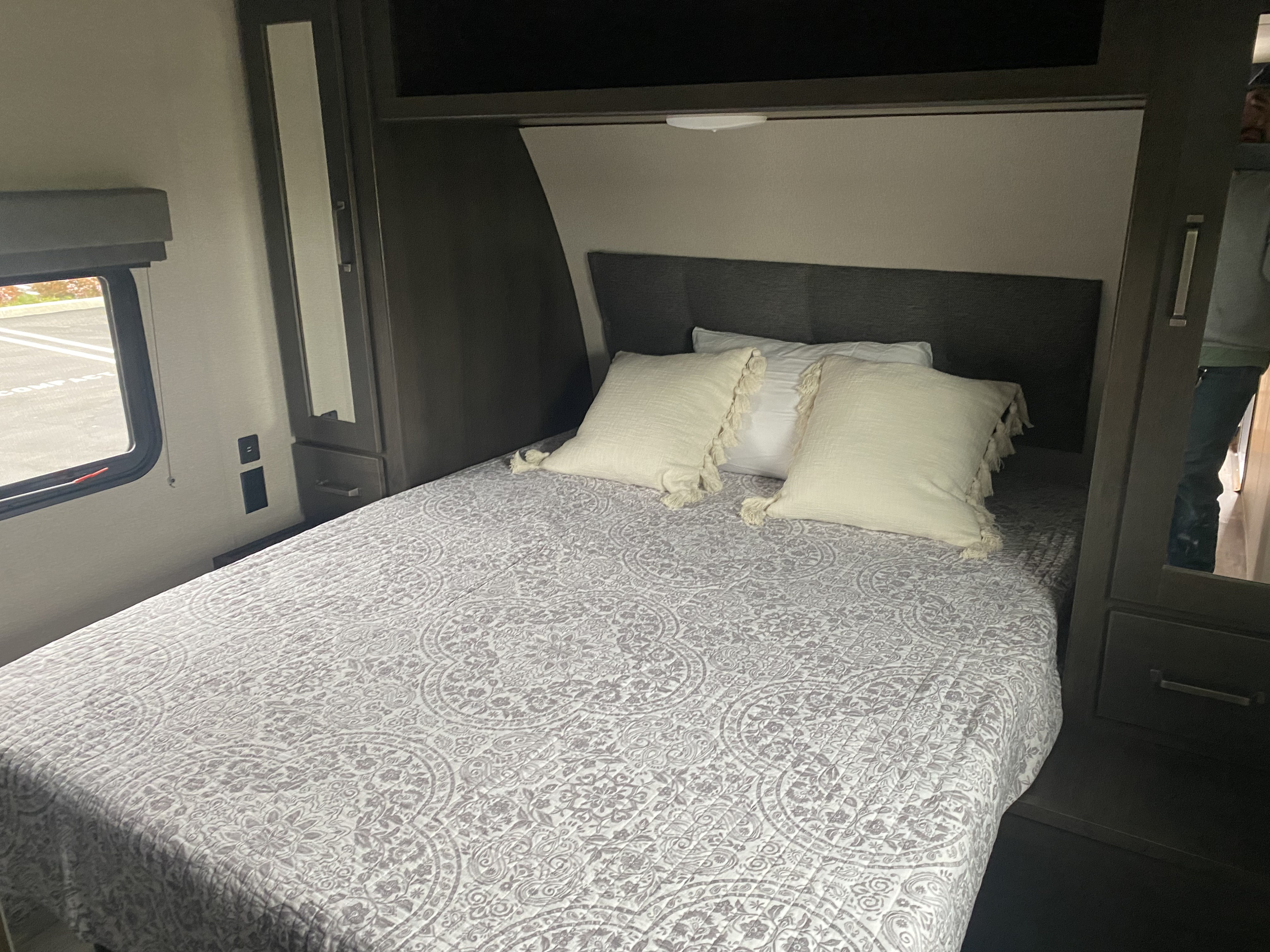 Cozy RV rental bedroom with a grey patterned quilt, white pillows, window view, and built-in storage on both sides.. Grand Design Transcend Xplor 2023