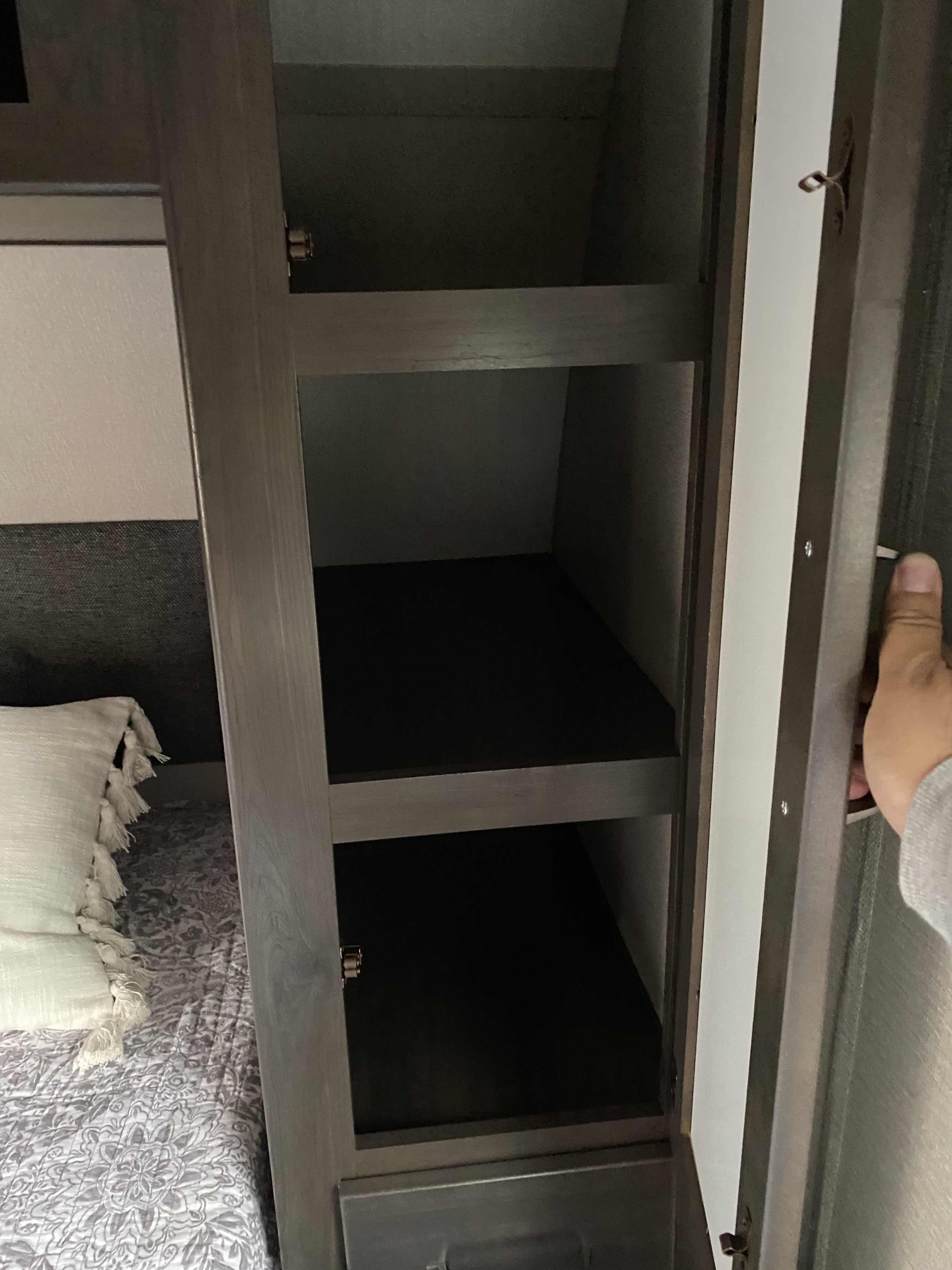 This rv rental bedroom features an open closet with multiple dark shelves, perfect for storage, alongside a patterned bed.. Grand Design Transcend Xplor 2023