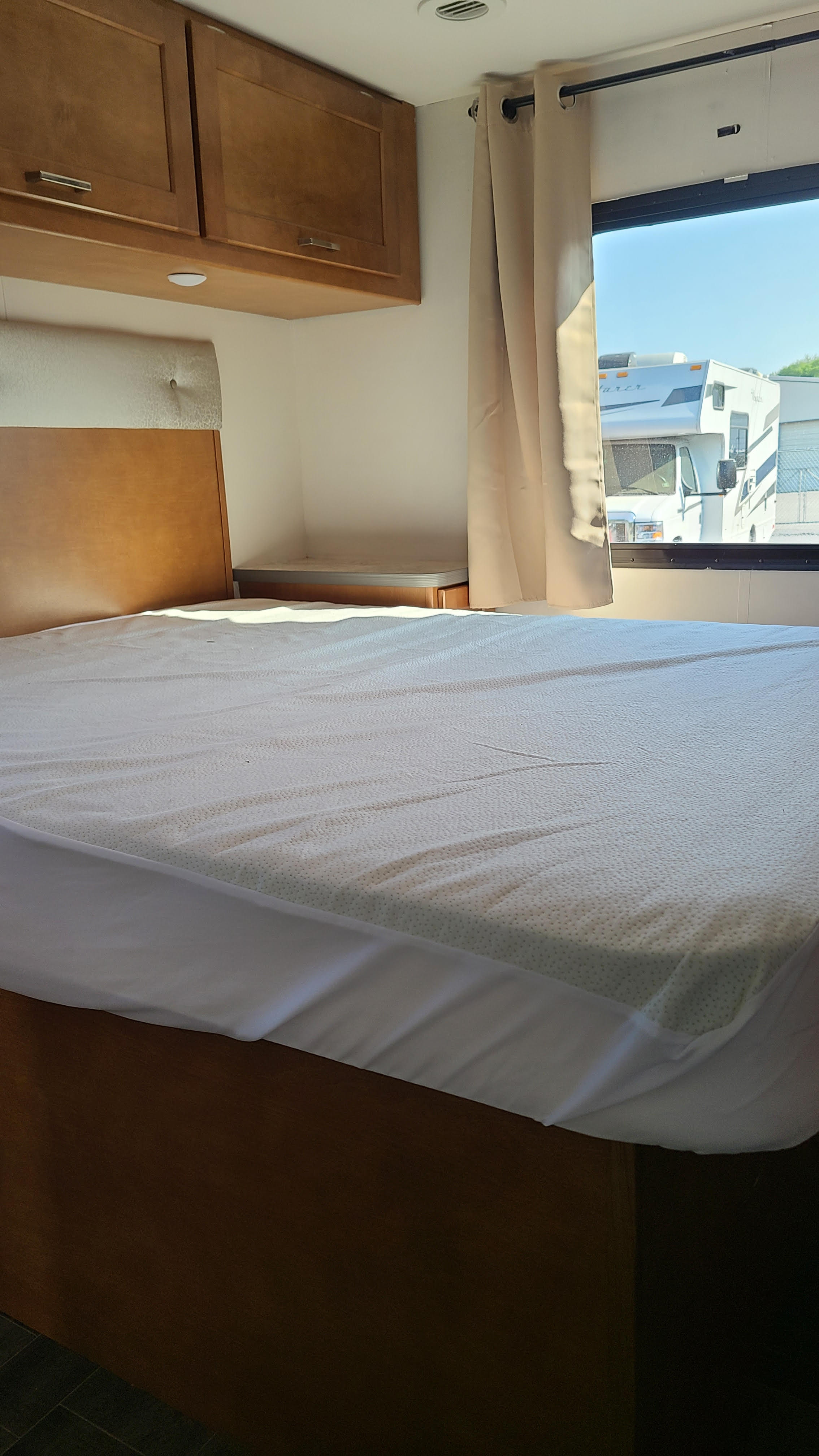 RV rental bedroom features a comfy bed with overhead cabinets & a window view of another RV, ensuring a cozy stay.. Winnebago Minnie Winnie 2019