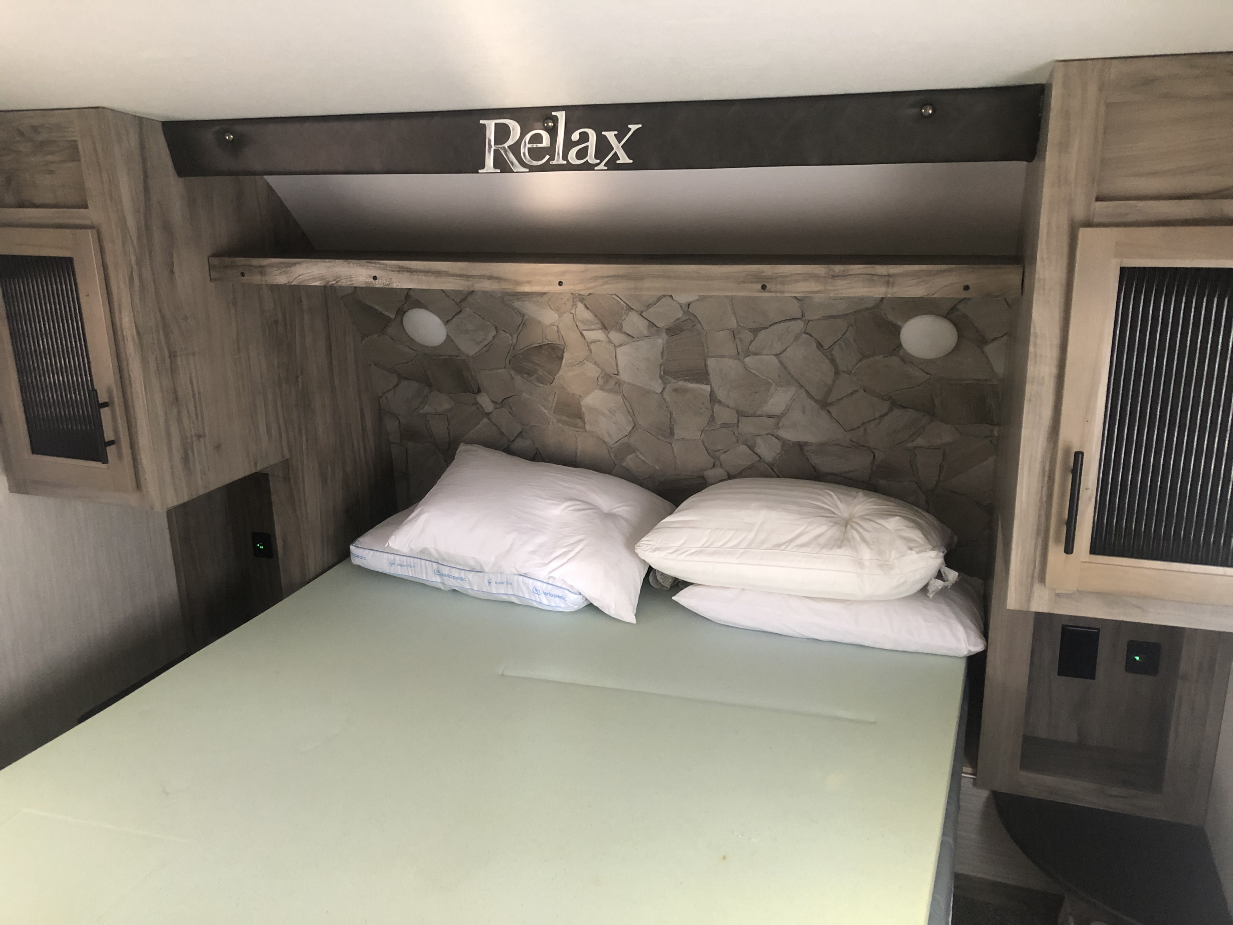 Cozy RV rental bedroom featuring a bed, pillows, "Relax" sign, and functional storage space.. Forest River Cherokee Arctic Wolf 2020