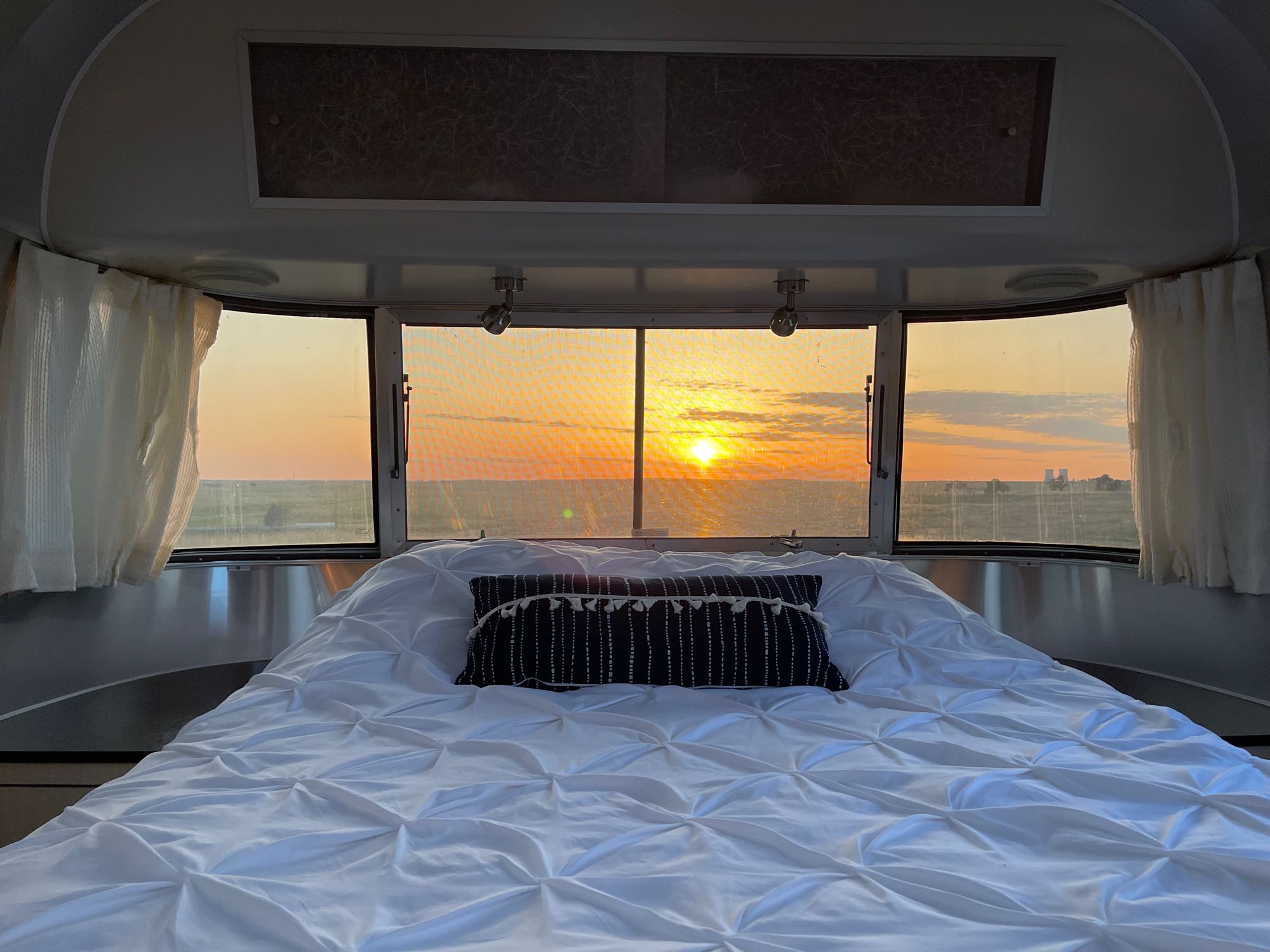 RV rental bedroom view: a white bed faces panoramic windows showcasing a vibrant sunset over an open landscape.. Airstream International 2011