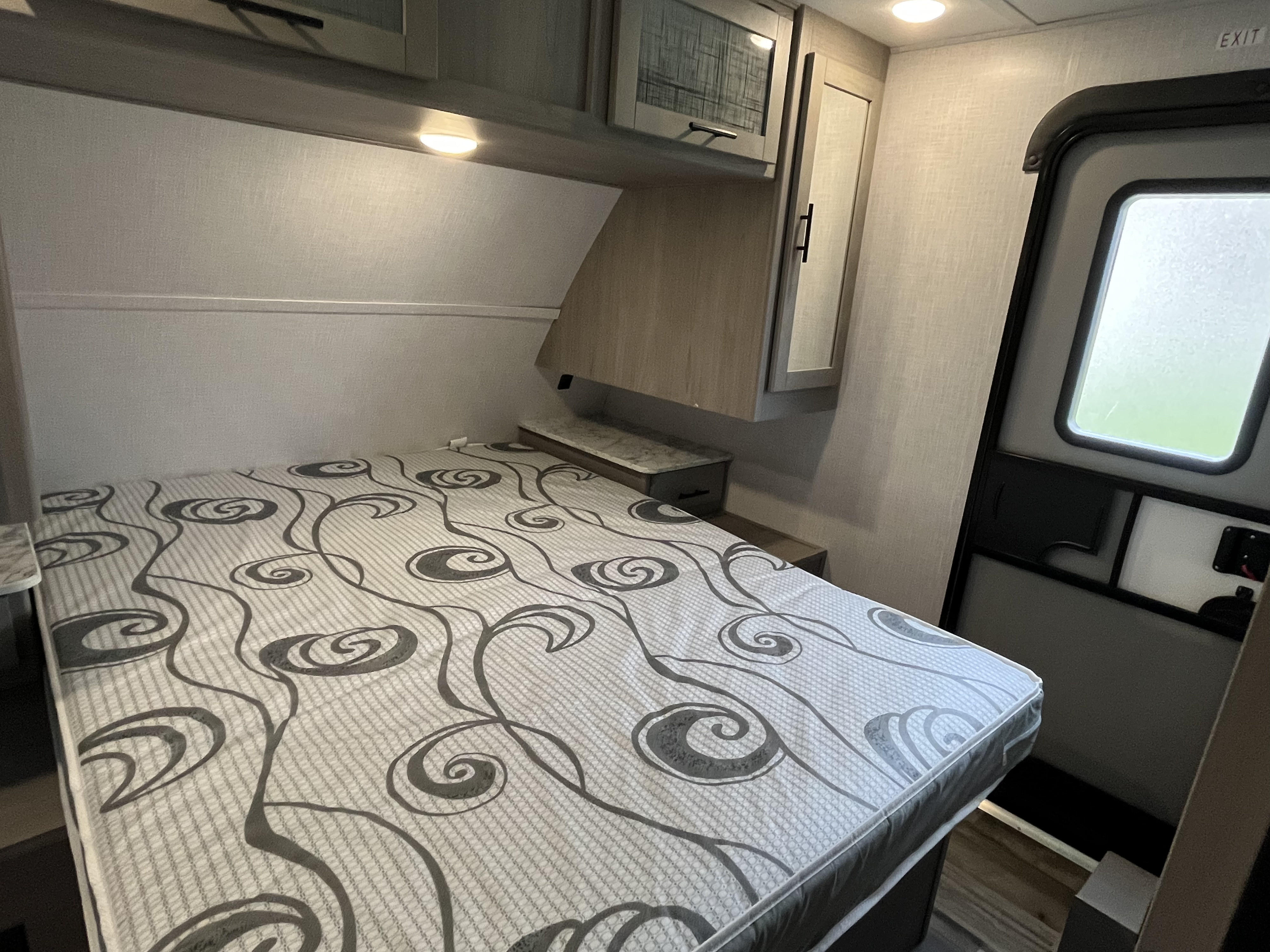 Comfortable RV rental bedroom with patterned mattress, overhead cabinets, under-cabinet lighting, and a side door.. Dutchmen Coleman Light 2022