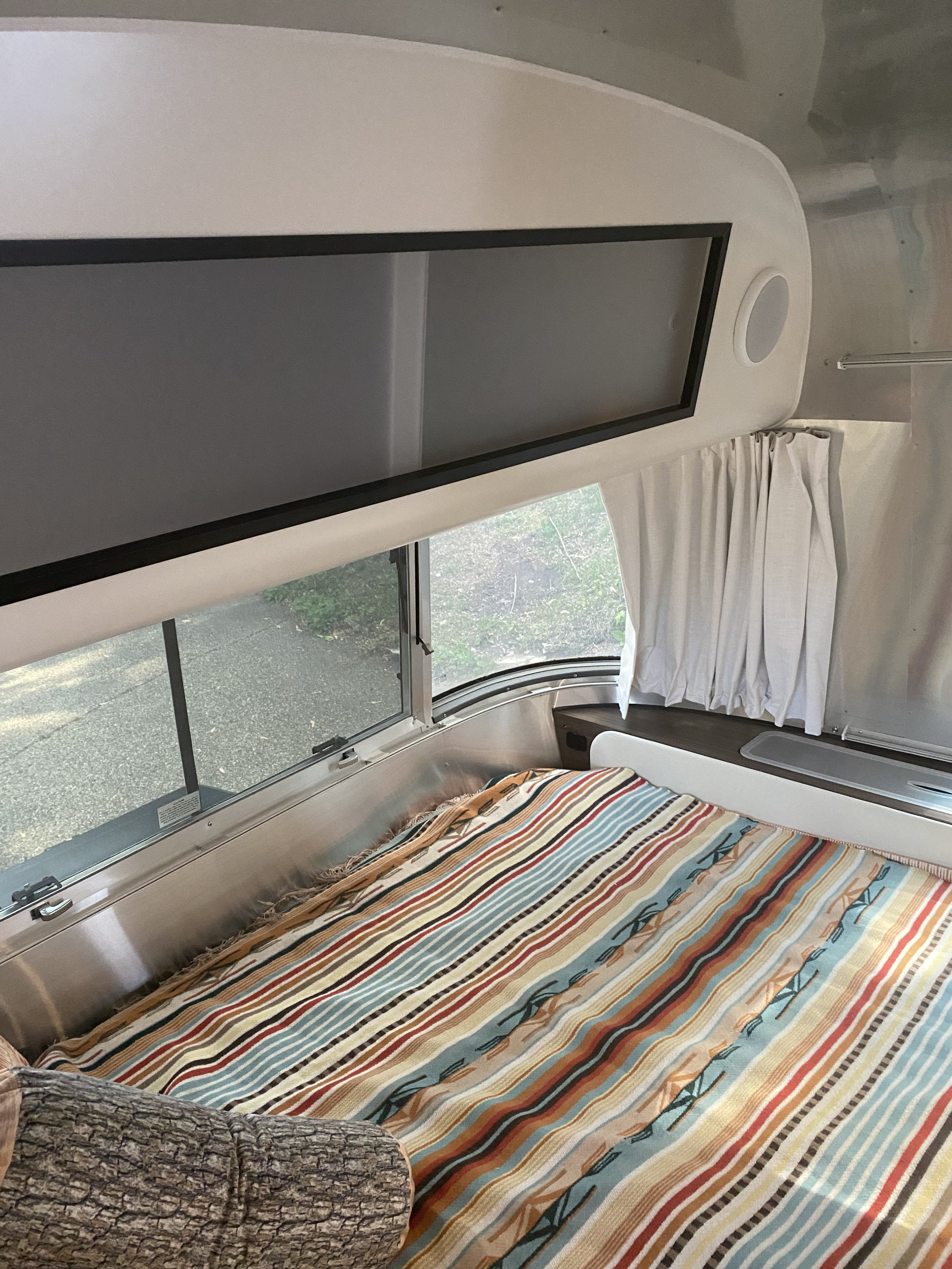 Interior of a modern Airstream RV rental: cozy bed with vibrant striped blanket, large window view, and overhead storage.. Airstream Caraval 2021