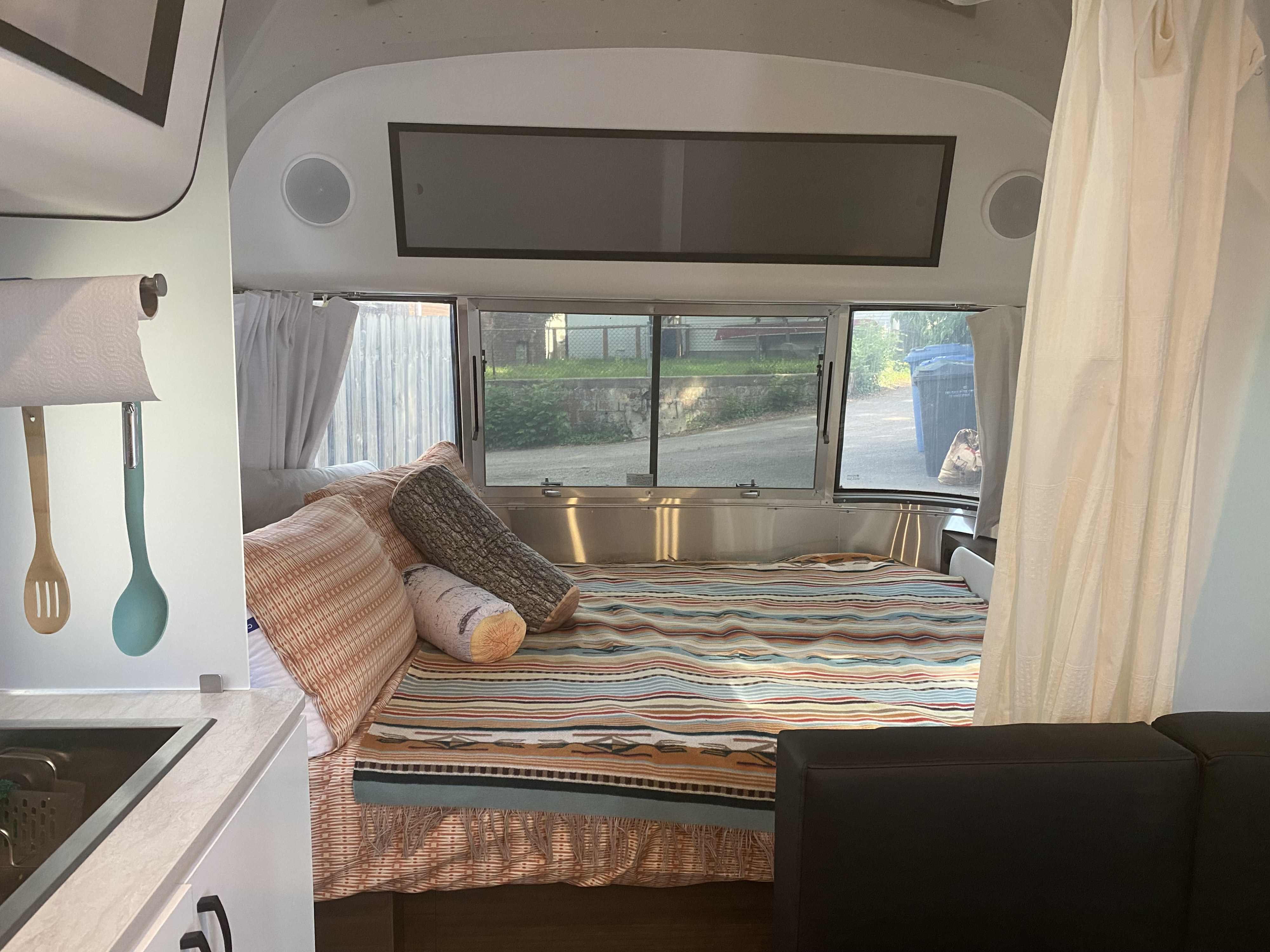 Inside a comfortable RV rental: a bed with unique log pillows, a kitchen corner, and windows showing an outdoor view.. Airstream Caraval 2021