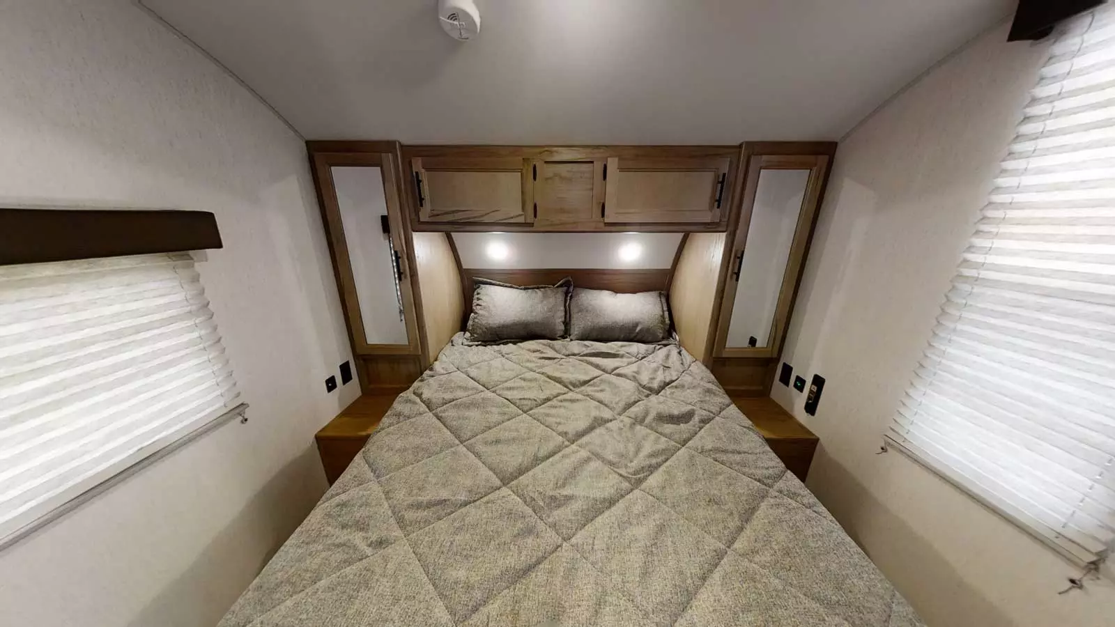 A full view of an RV rental bedroom with a quilted bed, overhead cabinets, and windows with blinds on both sides.. Forest River 23mk 2023