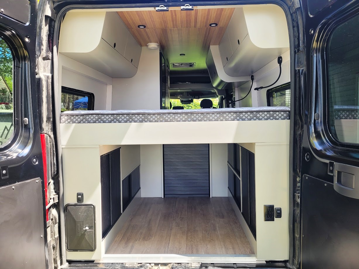 Modern RV rental interior with elevated bed & large storage space below. Clean design, wood floor & ceiling visible inside camper van.. Promaster 3500 EXT 2022