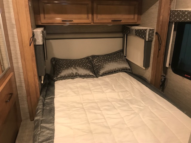 Cozy RV rental bedroom with a neatly made bed, grey pillows, overhead storage cabinets, and pull-down window shades.. Thor Quantum SE 2021