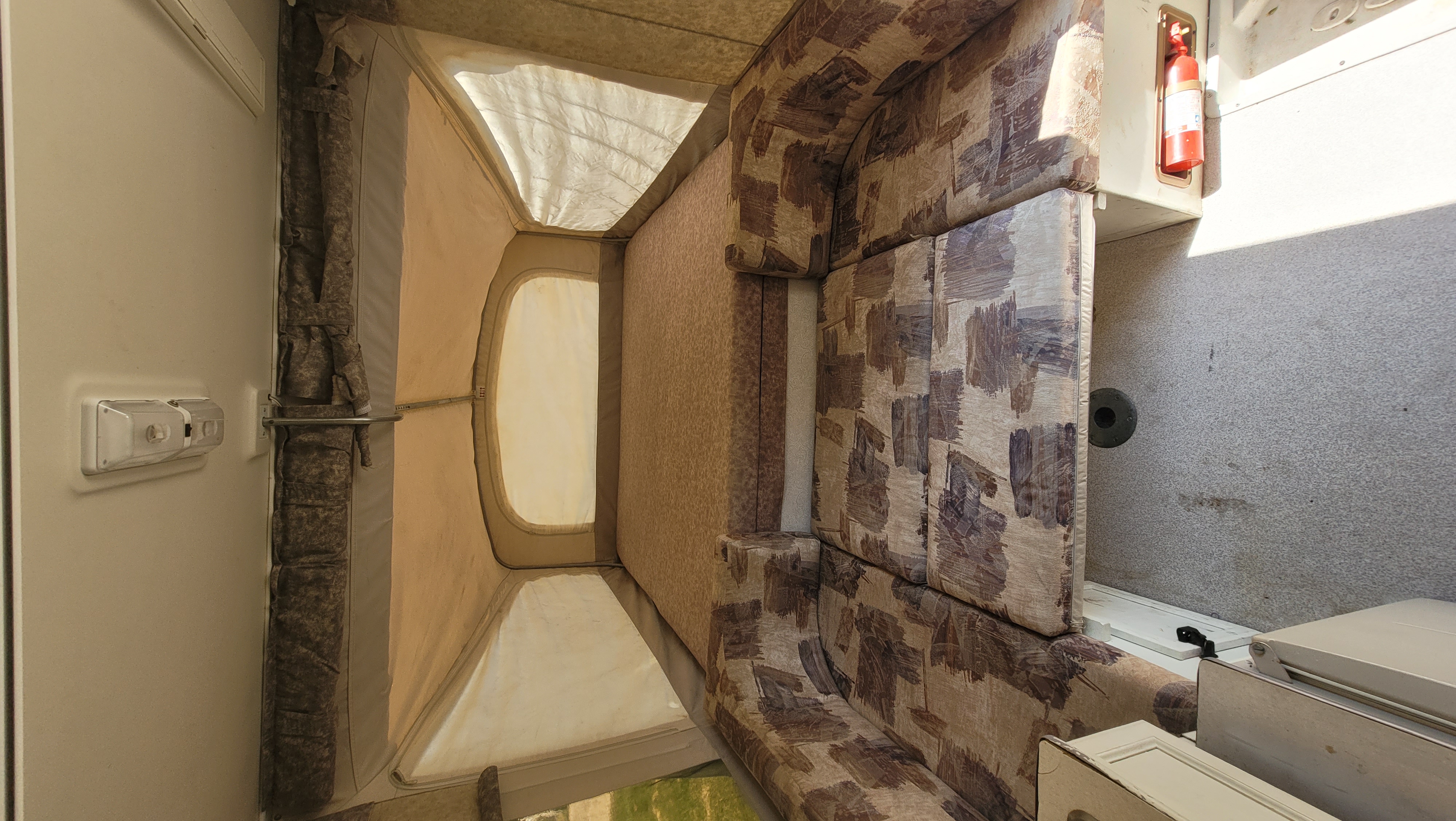 Interior of an RV rental featuring a cozy, patterned dinette area and a light-filled pop-out tent bed. A fire extinguisher is mounted.. Jayco Heritage 2000