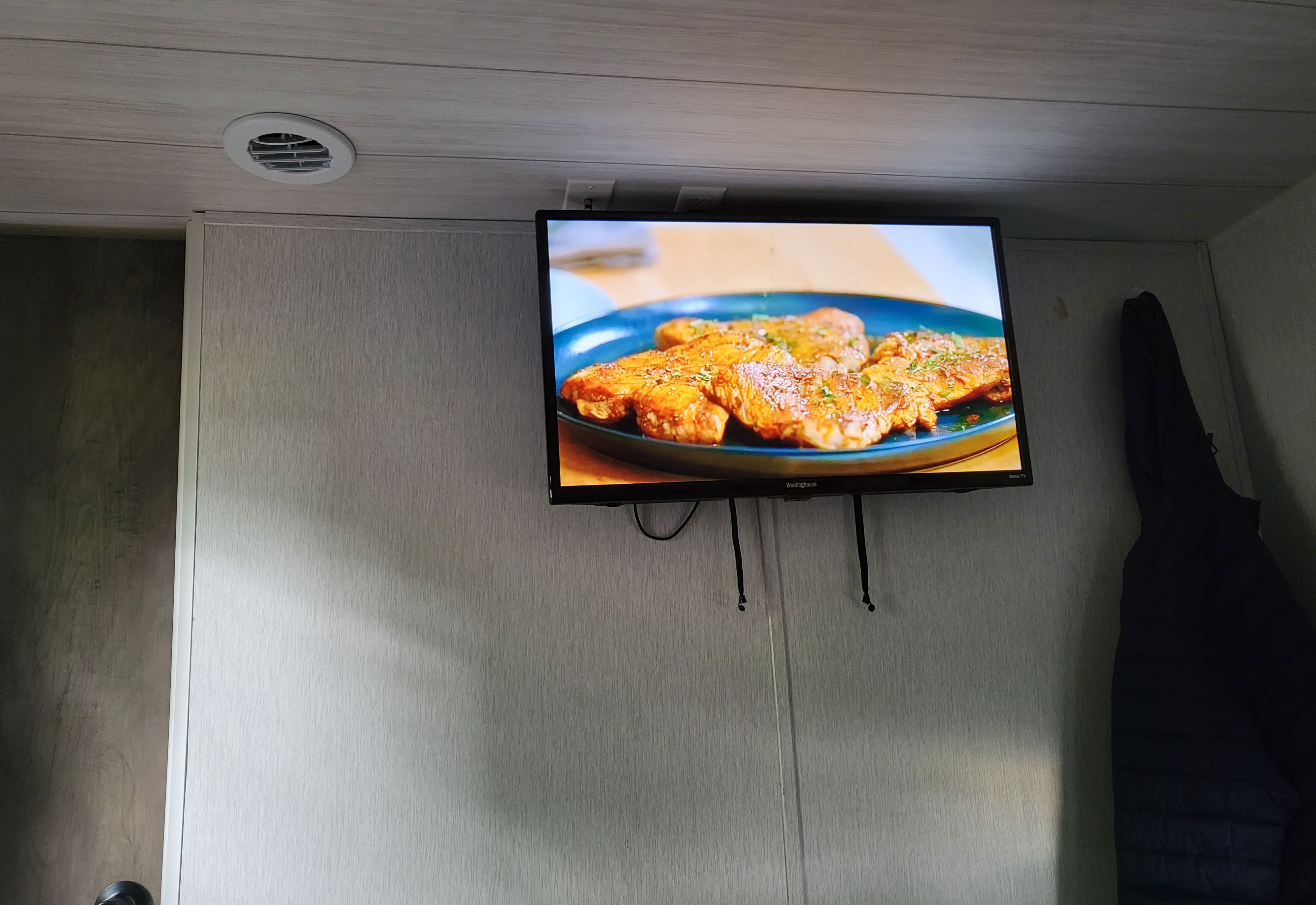 Wood-paneled RV rental interior with a TV displaying cooked food. A ceiling vent and hanging jacket are visible.. Forest River Wildwood X-Lite 2022