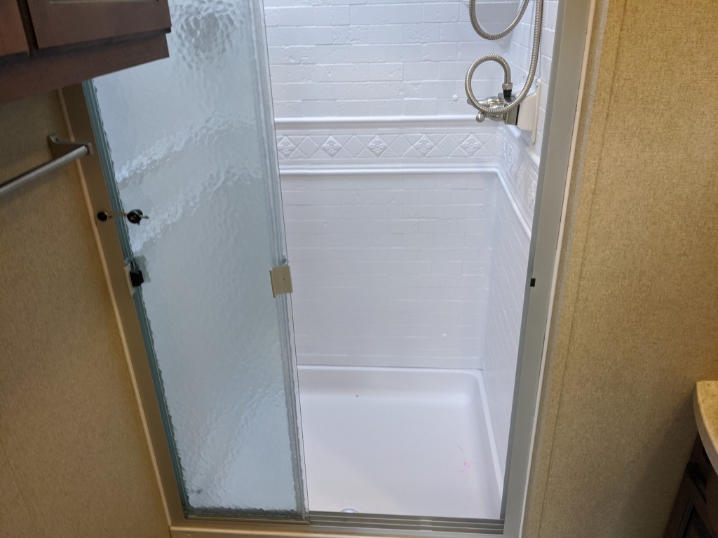 Bright, compact RV rental shower with white tiled walls, shower head, and clear glass sliding door.. Grand Design Reflection 2018