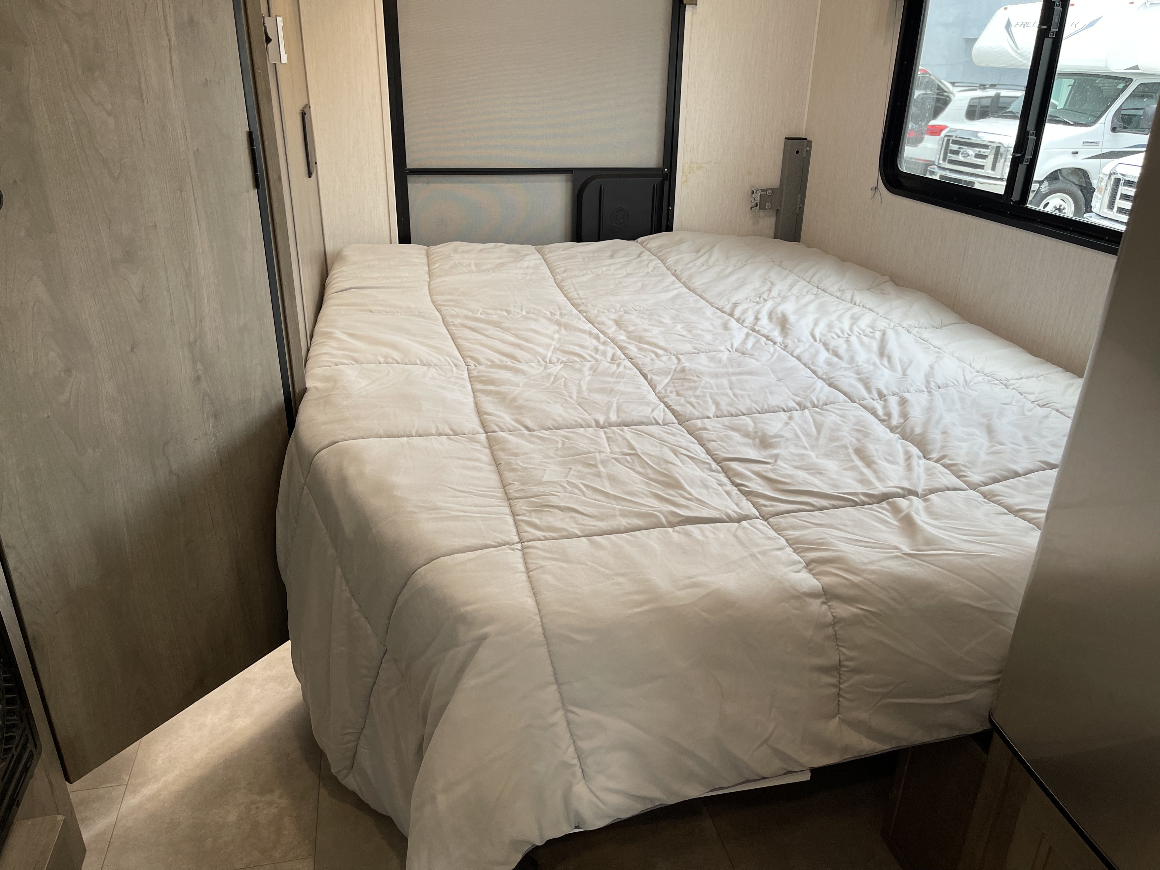 Relaxing RV rental bedroom with a comfy white quilted comforter, wood accents, and window looking out to other vehicles.. Coachmen Freelander 2021