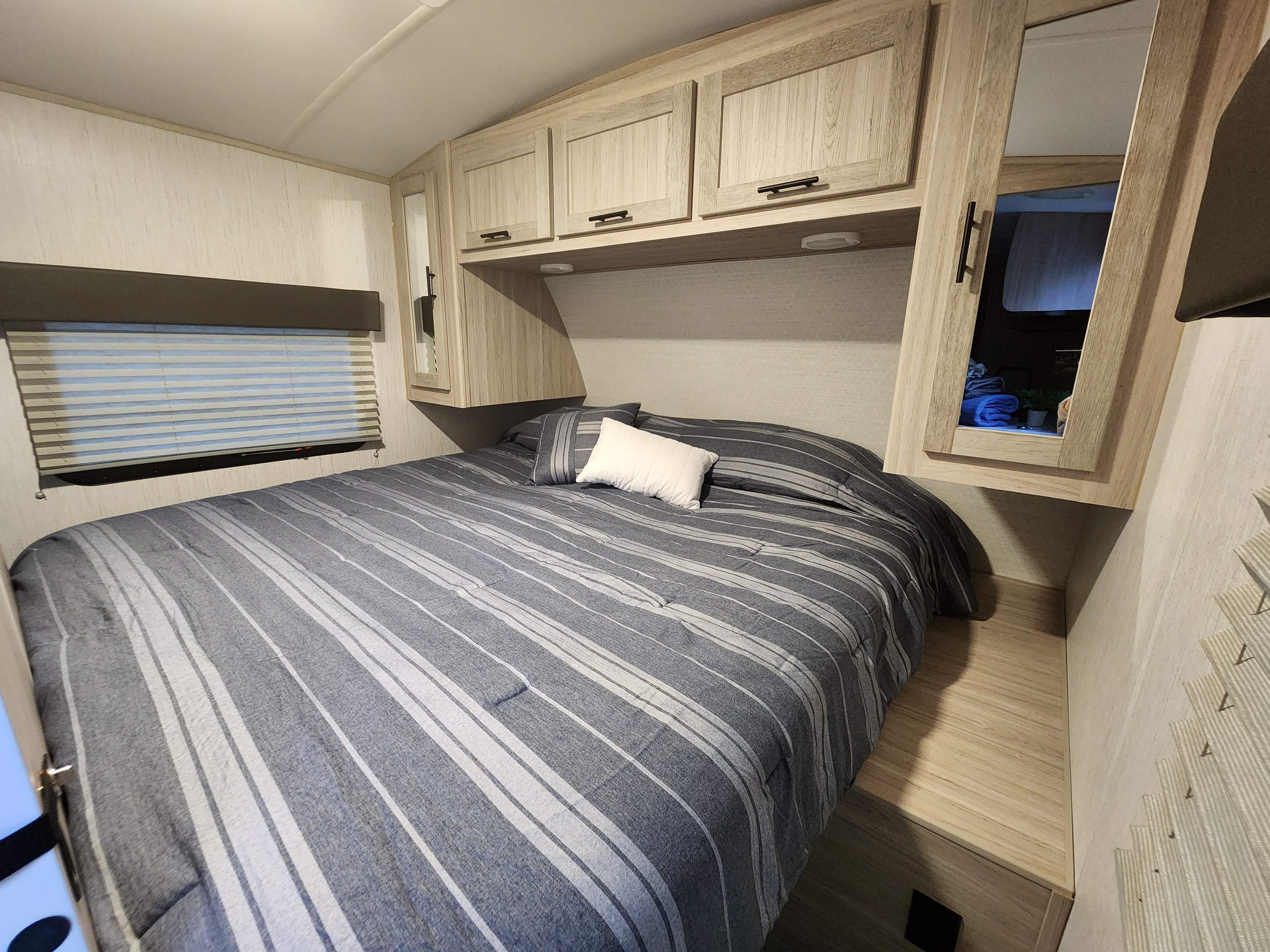 Light-toned RV rental bedroom with a striped bed, overhead cabinets, and window blinds, offering cozy interior comfort.. Heartland RVs Shadow Cruiser 2023