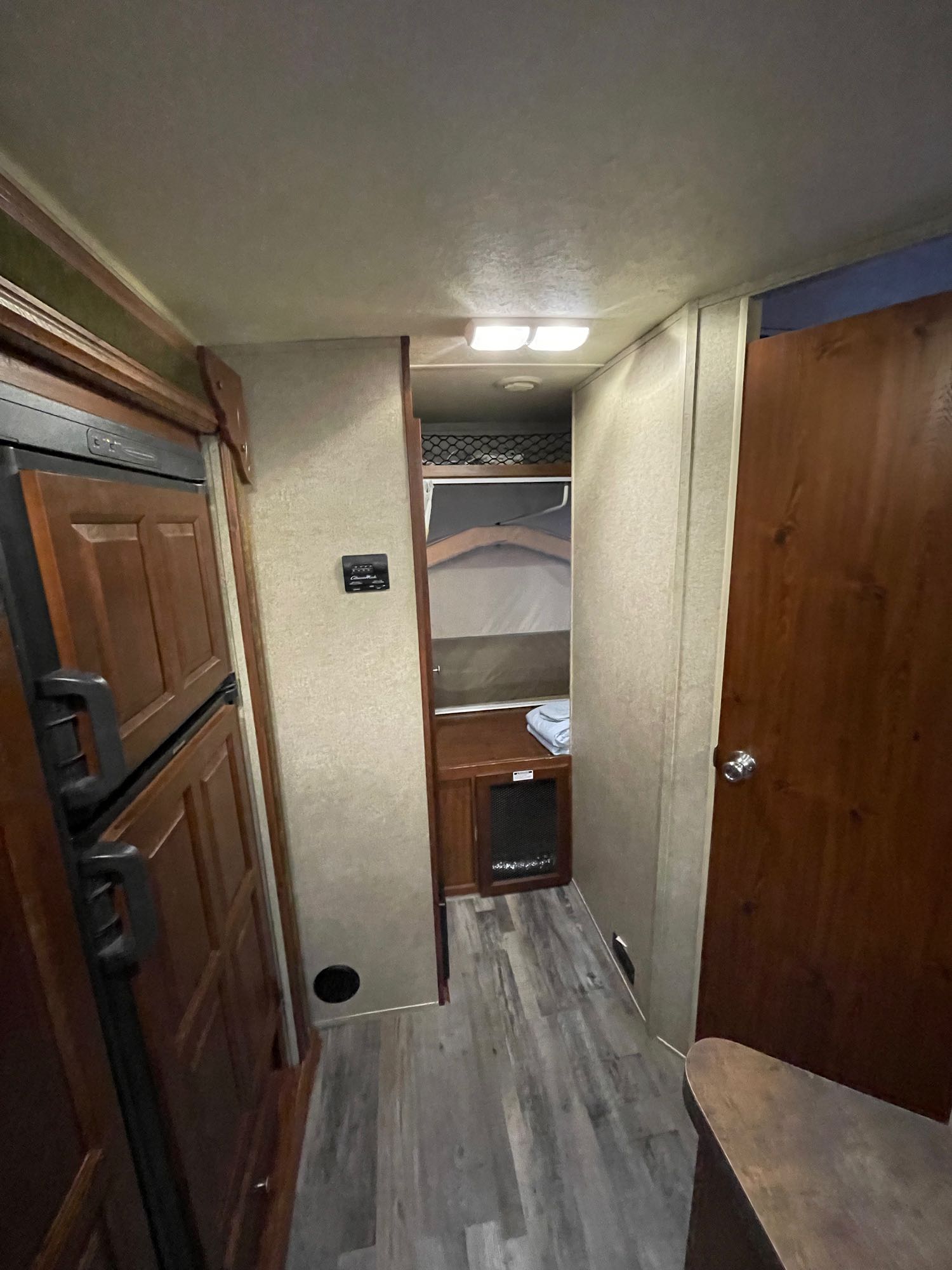 View of an RV rental interior with a wood-paneled fridge on left, a narrow hallway, a pop-out bed, and a wooden door.. Forest River Rockwood Roo 2014