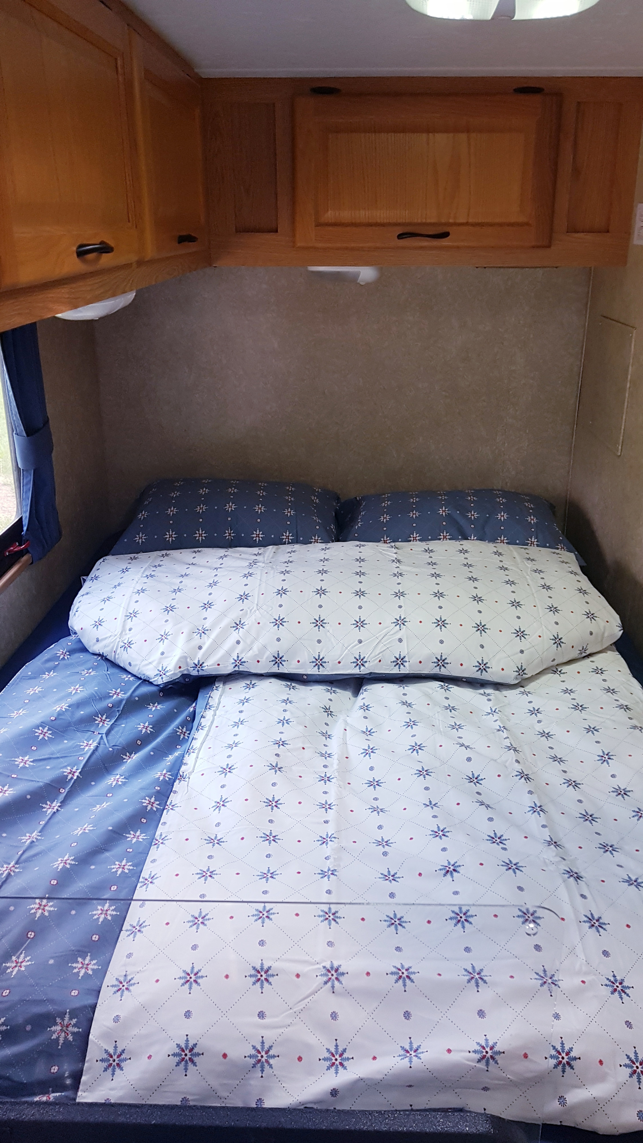 A cozy RV rental bed with snowflake-patterned blue and white bedding, and wood overhead cabinets.. Thor Majestic 2010