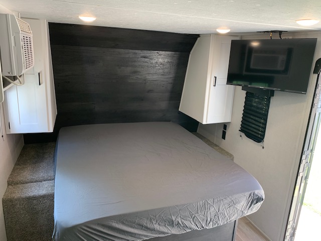 Cozy RV rental bedroom with grey bed, dark headboard, white cabinets, mounted TV, AC unit, and access steps.. Keystone RV Hideout 2021