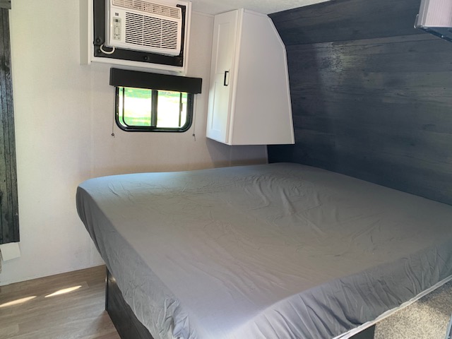 Cozy RV rental bedroom with gray bed, AC unit, and window. Overhead cabinet visible. Ready for your adventure.. Keystone RV Hideout 2021