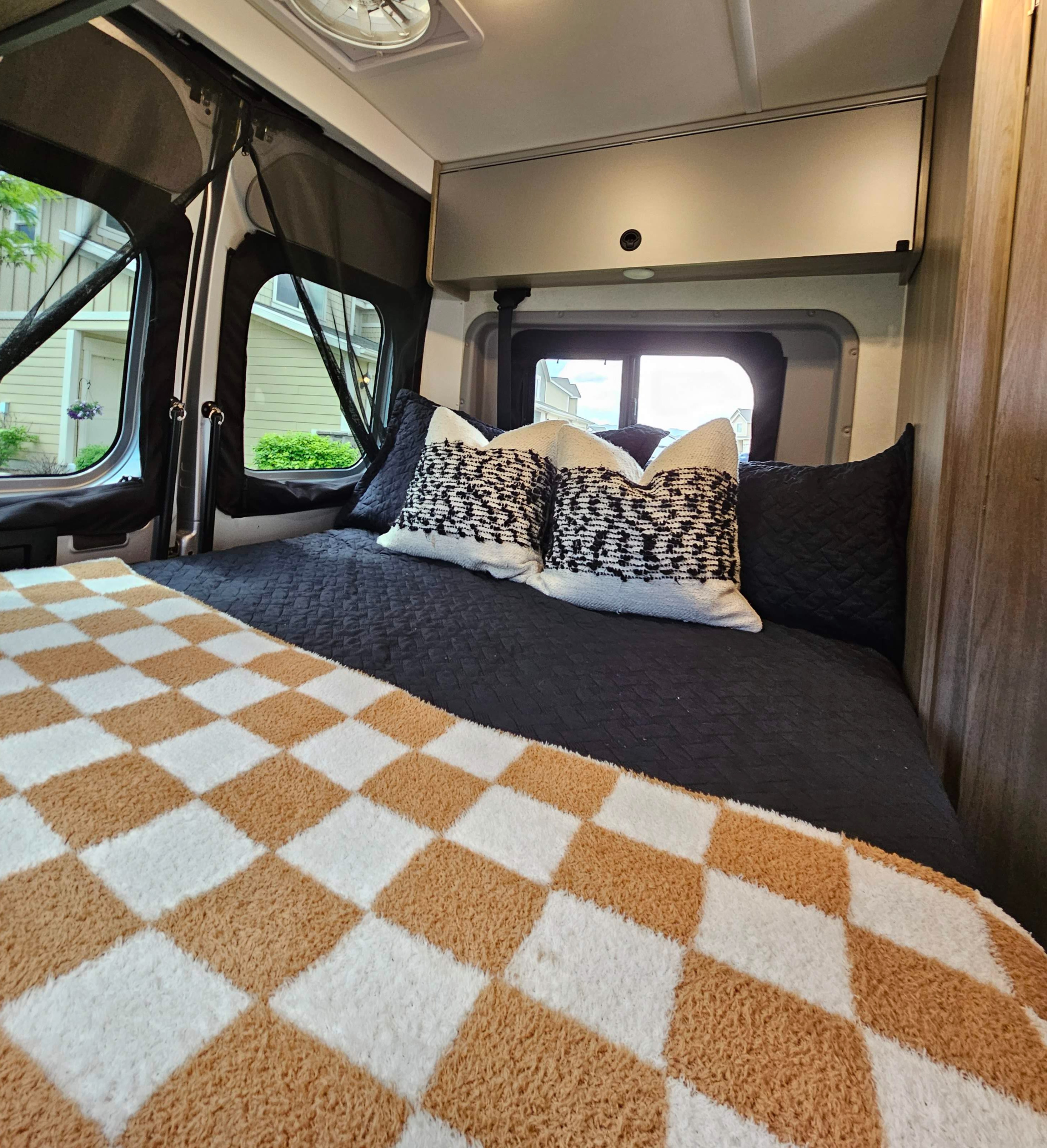 Inside a cozy RV rental: a comfy bed with stylish pillows, checkered throw, and serene window views.. Winnebago Solis 2022